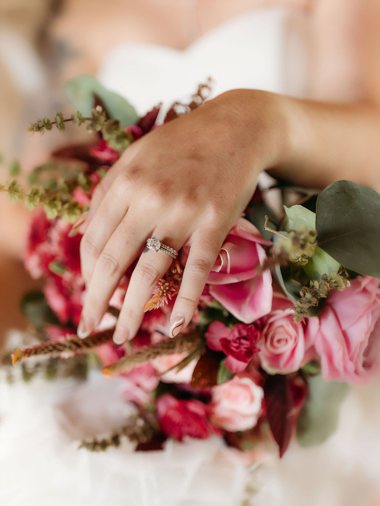 Bride displaying diamond engagement ring on hand resting over lush pink and red rose wedding bouquet.