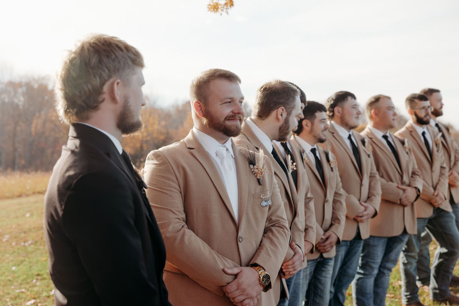 Groom and groomsmen in tan suits lined up outdoors at a rustic fall wedding ceremony.