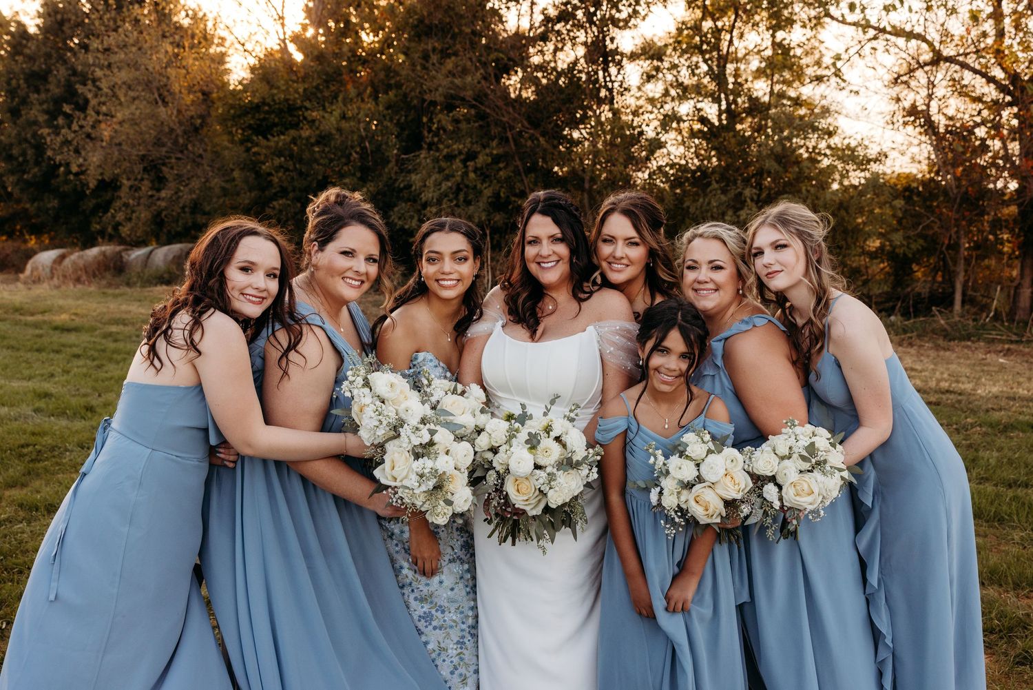 Bride and six bridesmaids in dusty blue gowns holding white bouquets pose together at golden hour outdoor wedding.