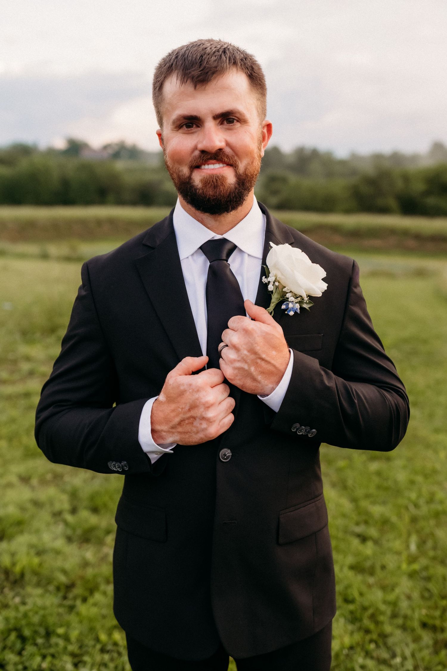 Smiling bearded groom in black suit and tie with white rose boutonniere posing in green field.