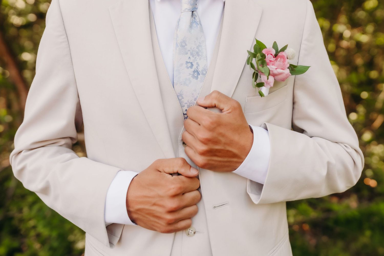 Groom in beige suit adjusting jacket with floral boutonniere and blue tie at wedding ceremony.