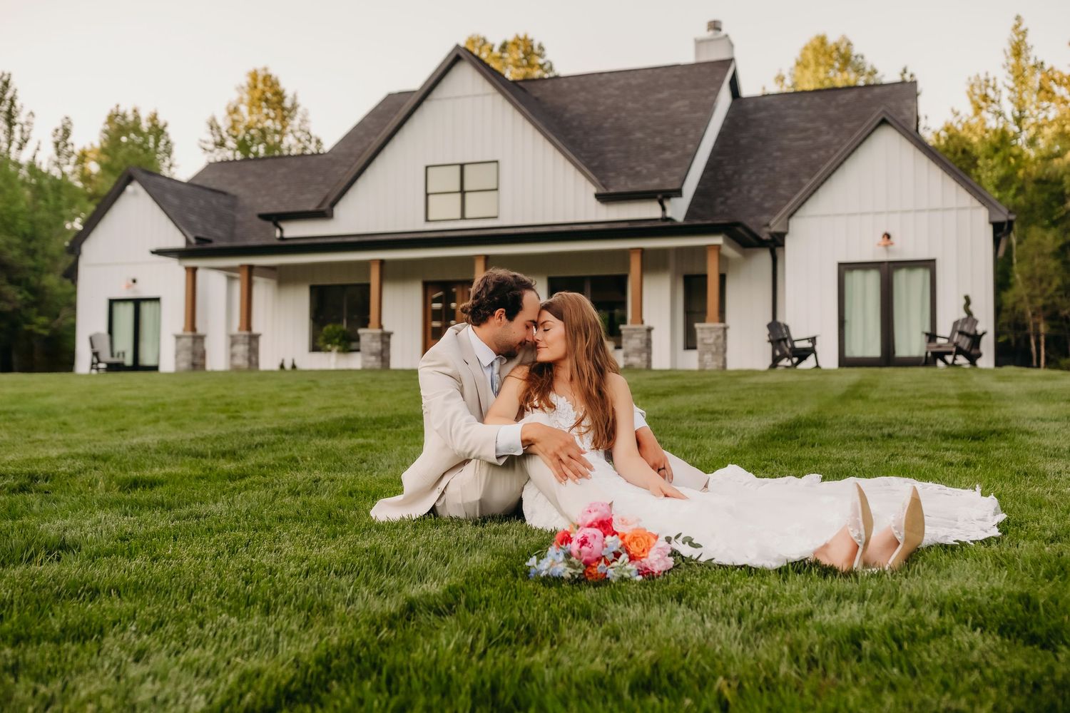 Bride and groom sitting on lush green lawn kissing in front of white farmhouse wedding venue.