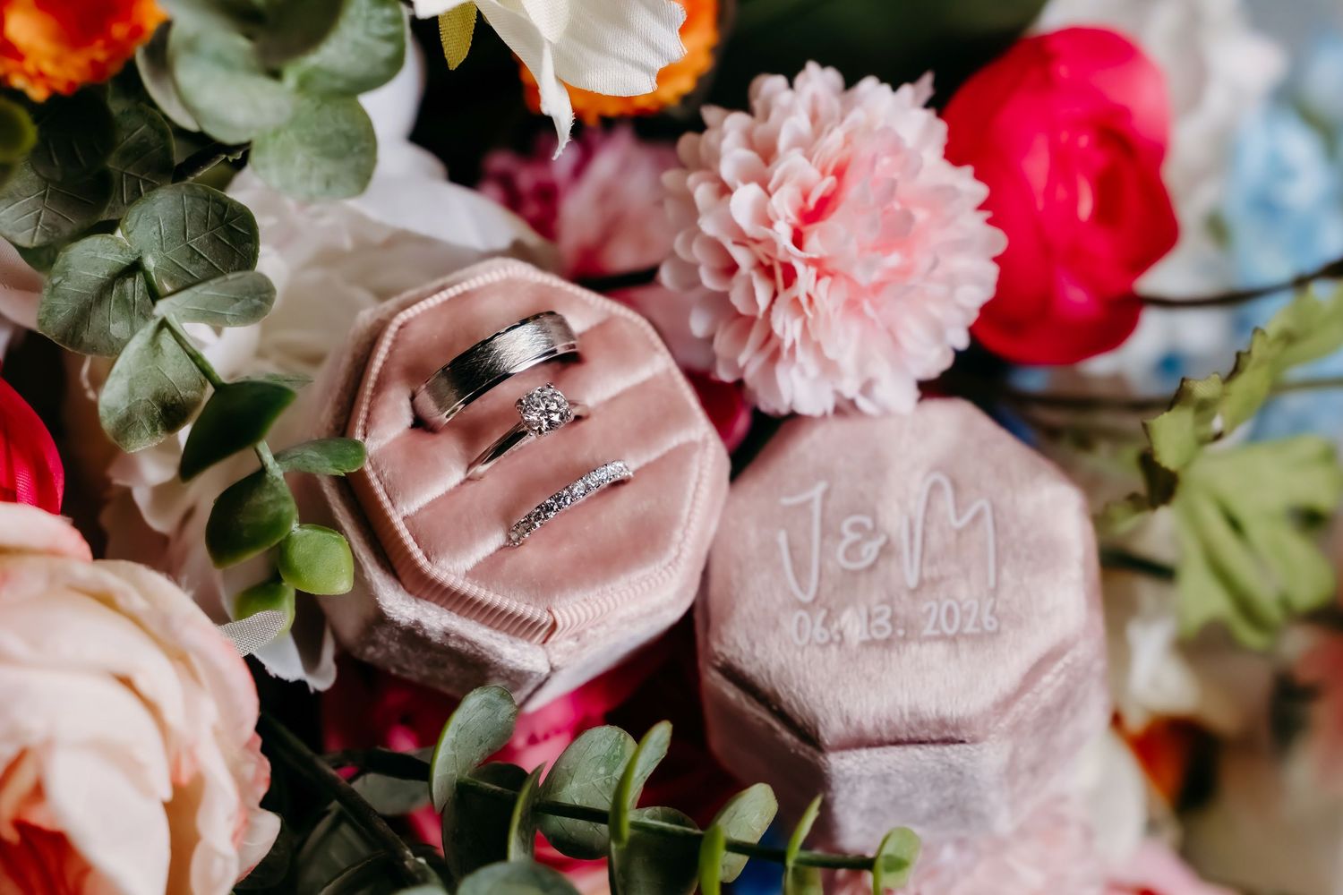 Wedding rings displayed in a pink velvet hexagonal box monogrammed J&M, surrounded by colorful floral arrangements.