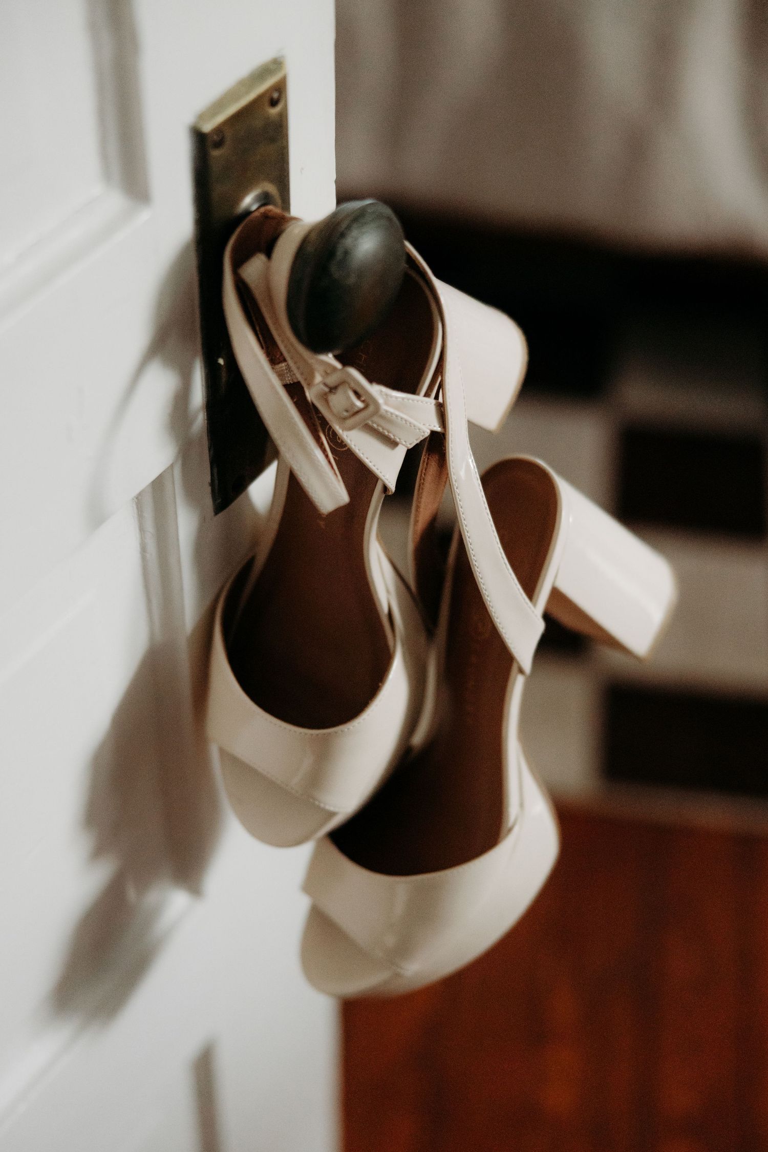 Elegant white block heel sandals with ankle straps hanging on a door knob, perfect bridal wedding shoes.