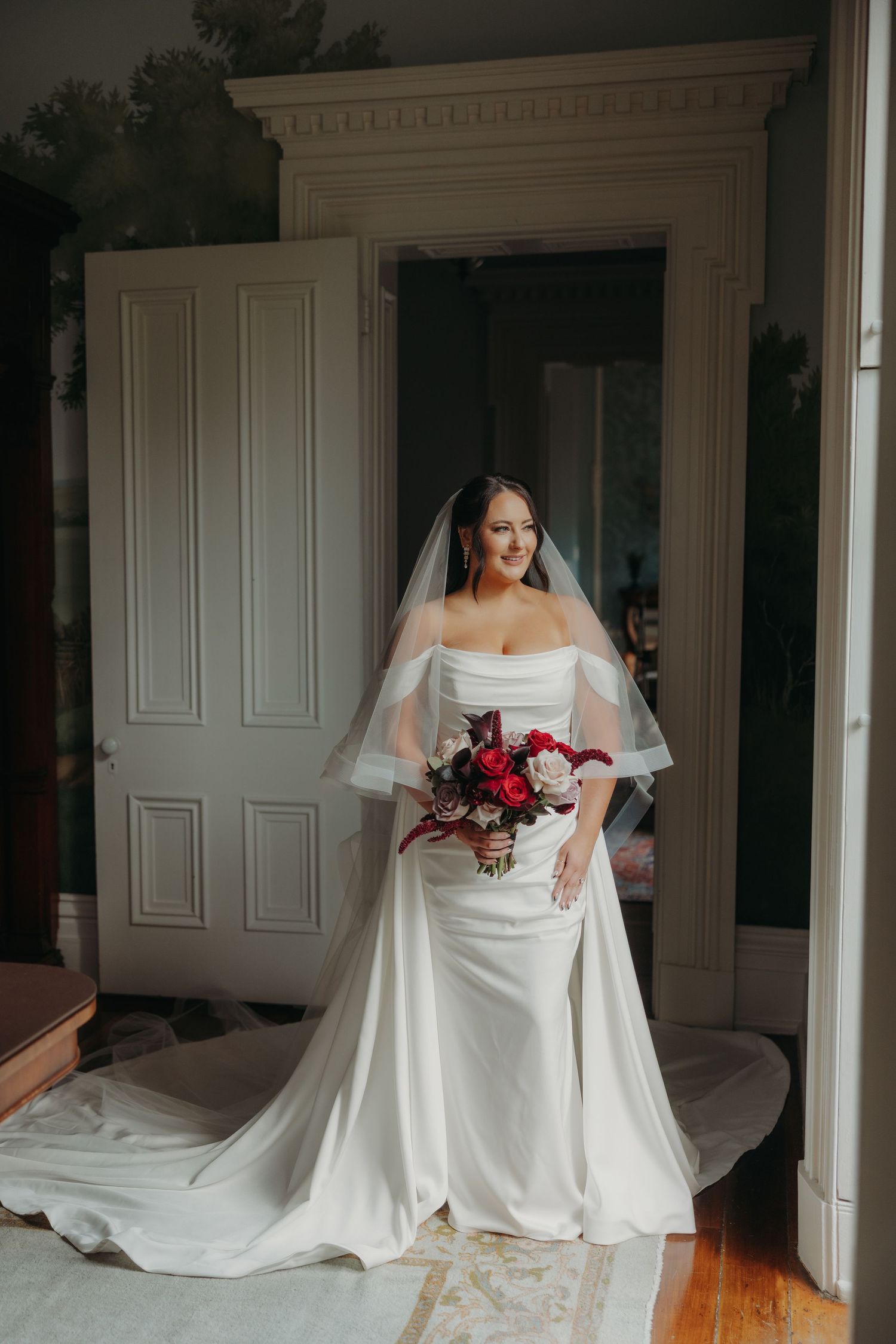 Bride in strapless white gown with veil holding red and white bouquet standing in elegant doorway.