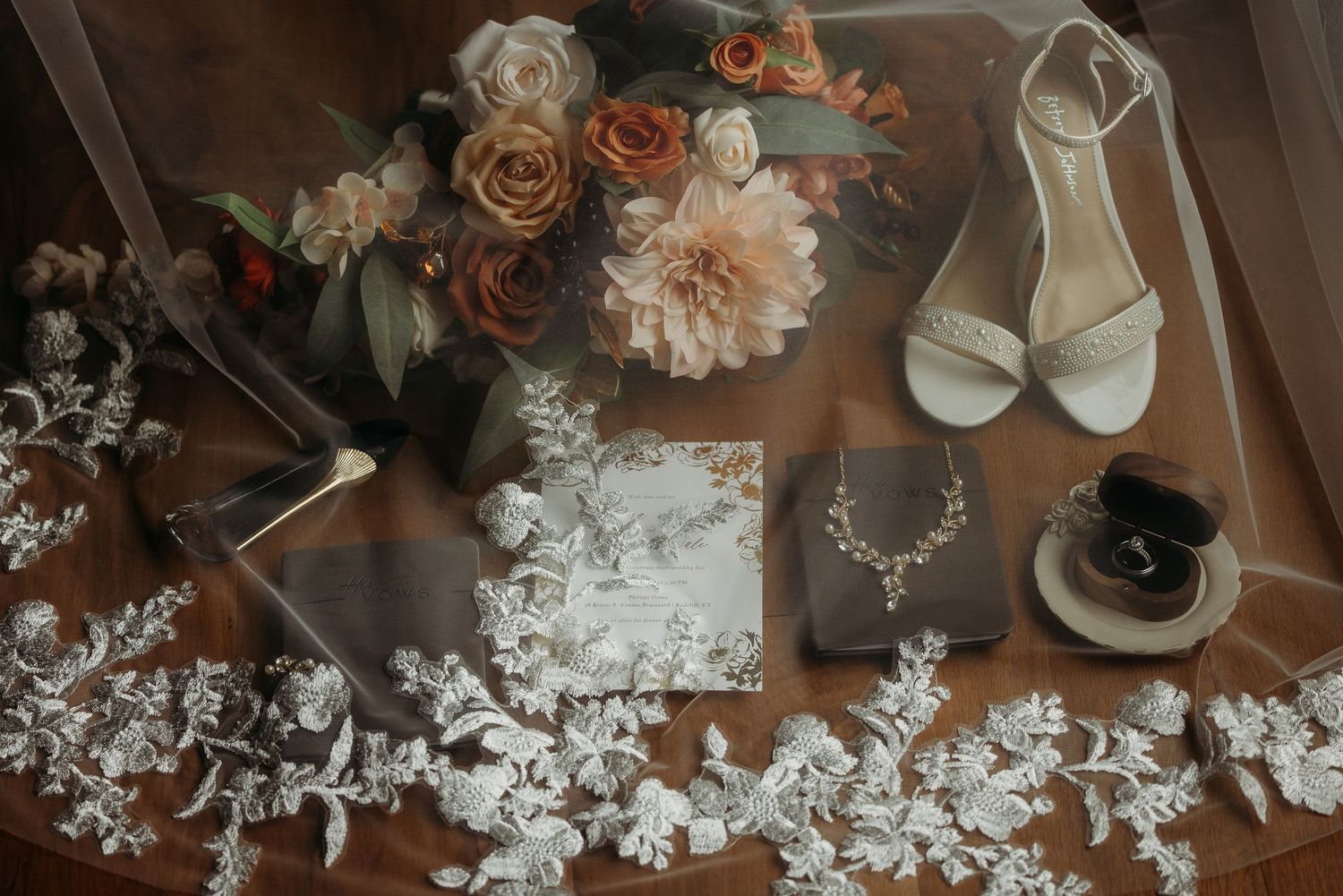 Elegant wedding flat lay featuring bridal shoes, floral bouquet, lace veil, jewelry, and accessories on a wooden surface.