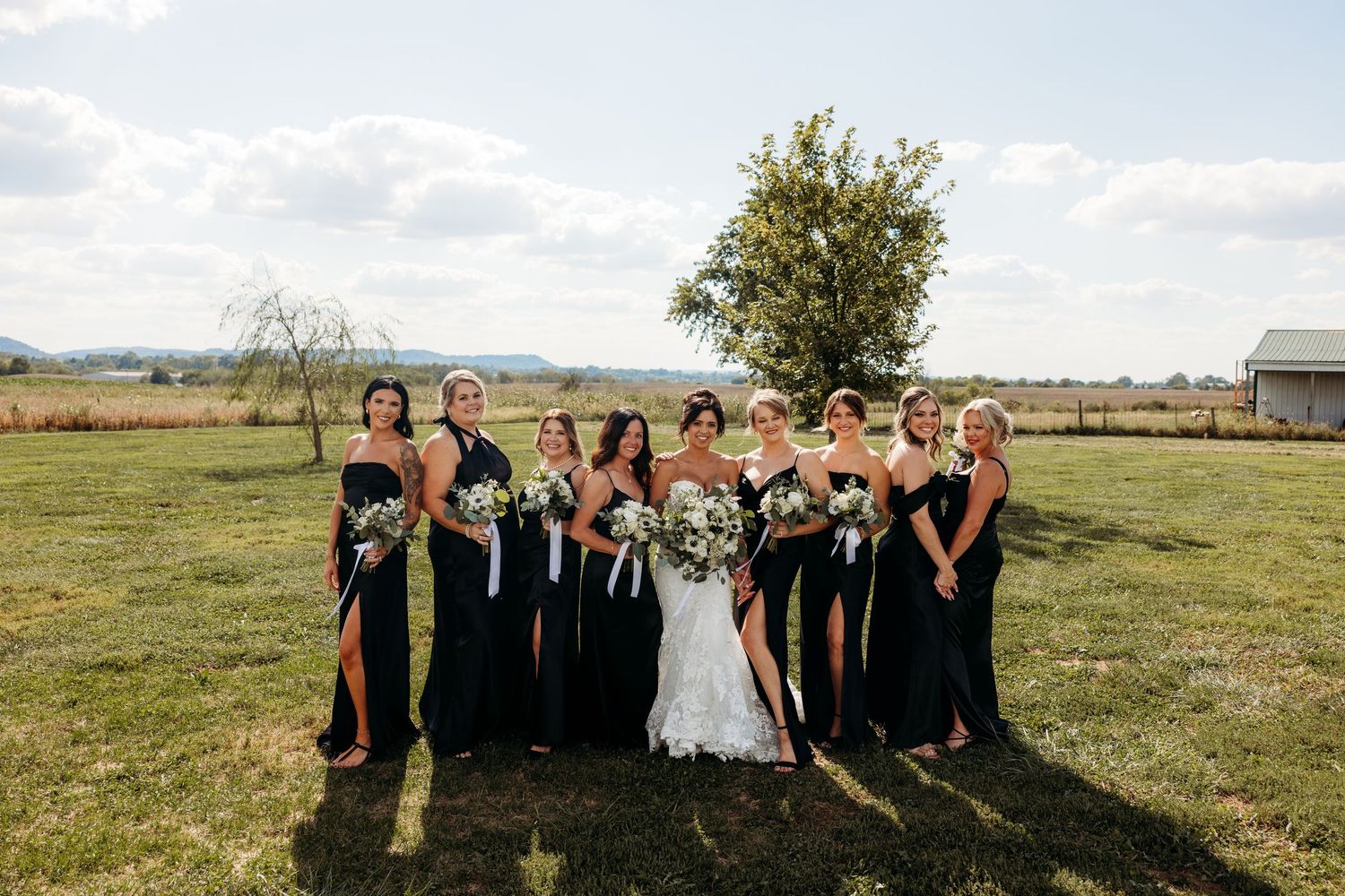 Bride and bridesmaids in black dresses posing together in a sunny green field on a beautiful wedding day.