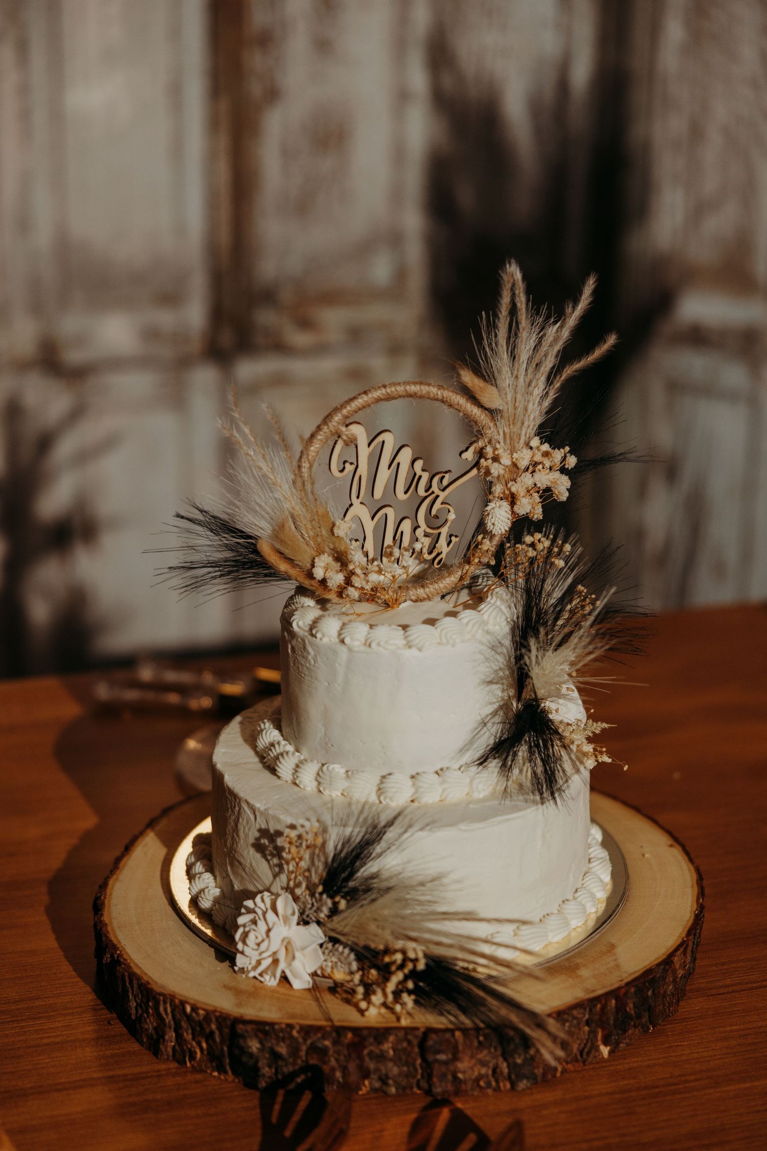Two-tier white wedding cake with boho pampas grass and feather decor topped with Mr & Mrs sign on wood slice.