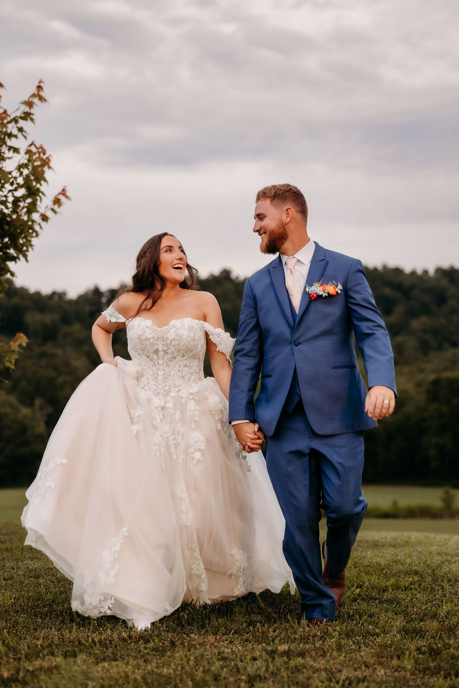 A joyful bride in an off-shoulder lace gown and groom in a blue suit hold hands while walking outdoors on their wedding day.