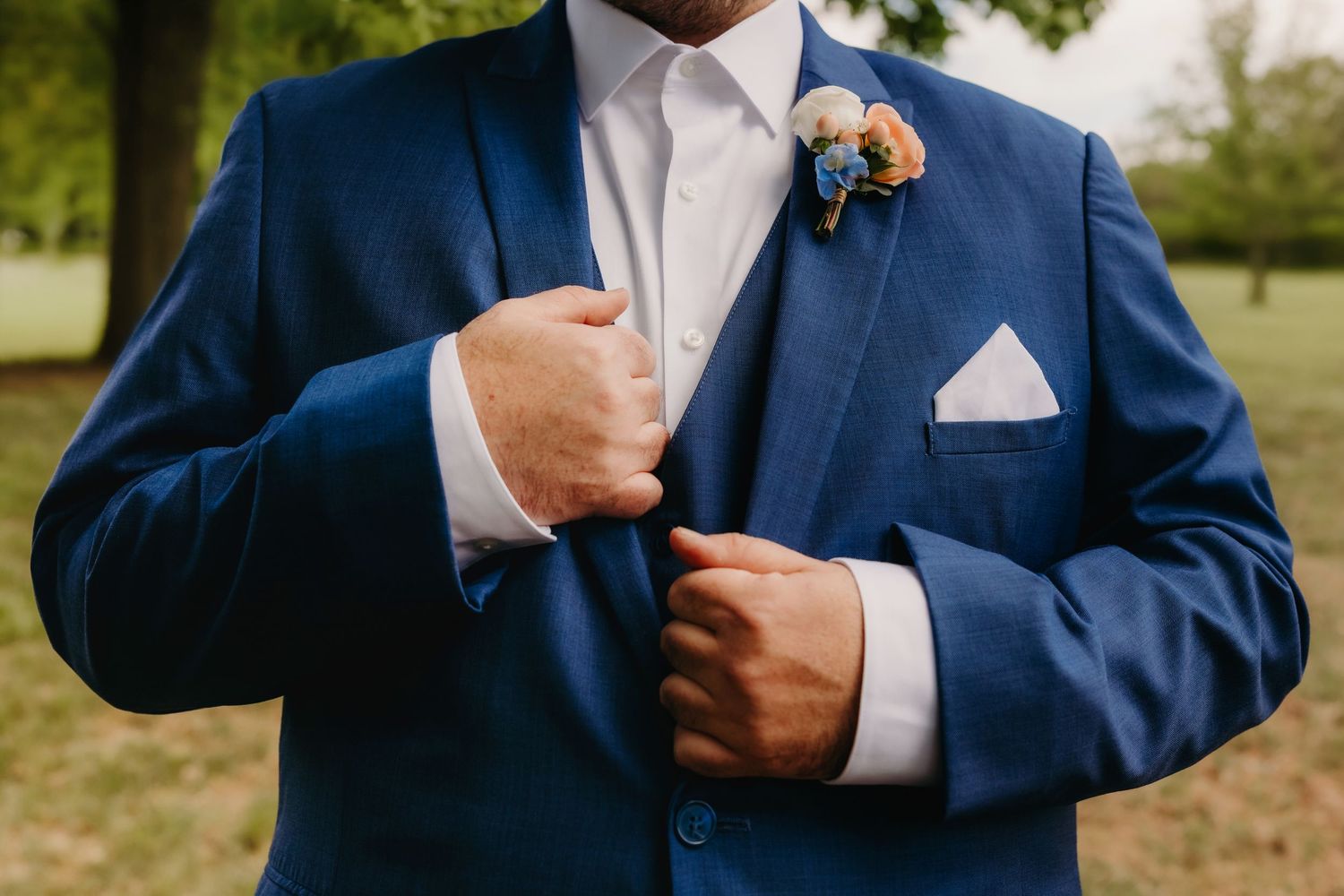 Groom in blue suit adjusting jacket with floral boutonniere and white pocket square outdoors.
