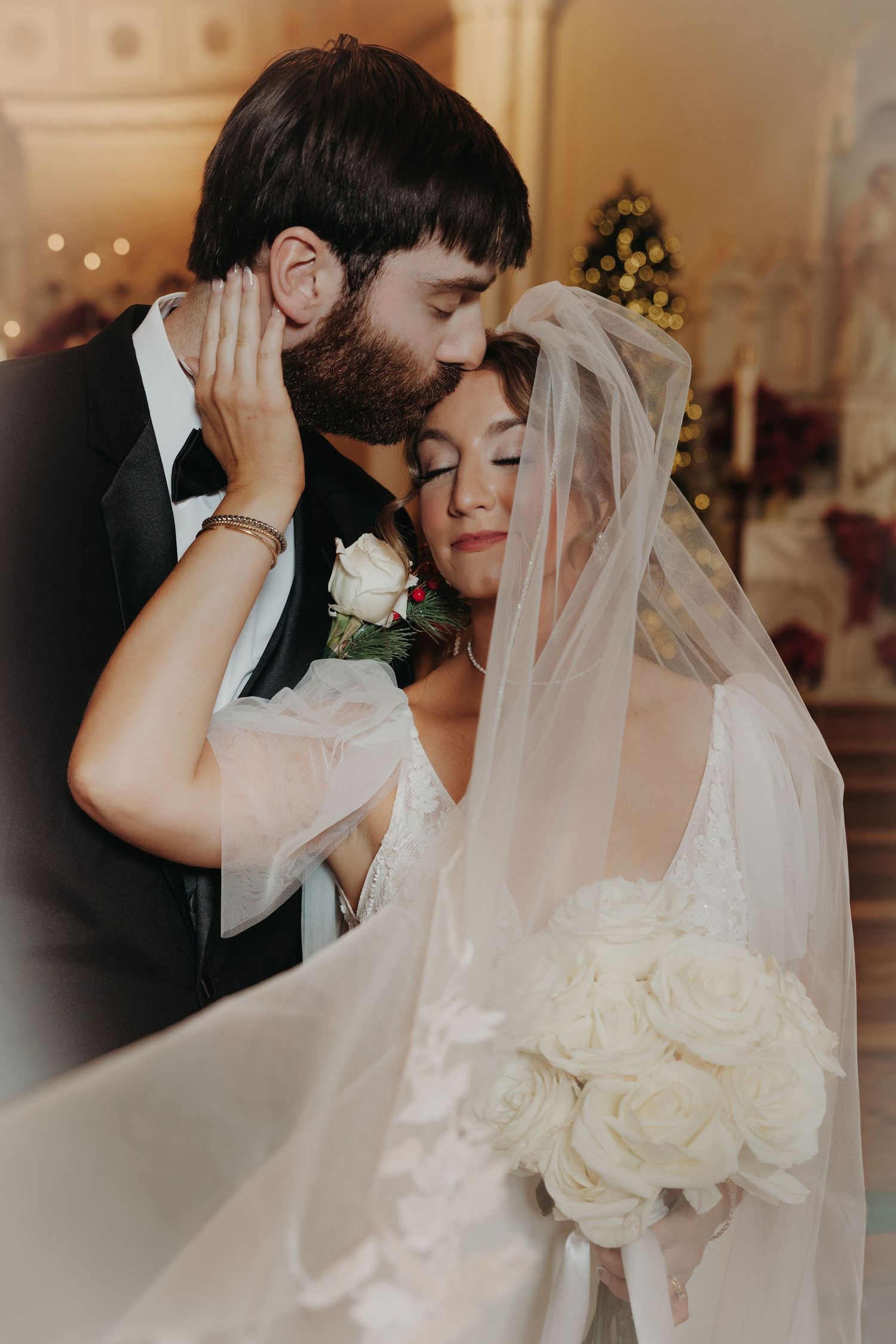 Groom tenderly kisses brides forehead as she holds white rose bouquet inside decorated Christmas church.