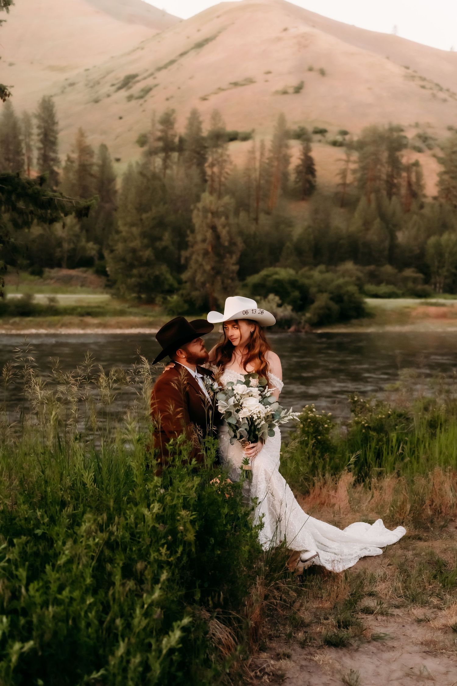 Bride and groom in cowboy hats share a romantic moment by a river with mountain backdrop at their western wedding.