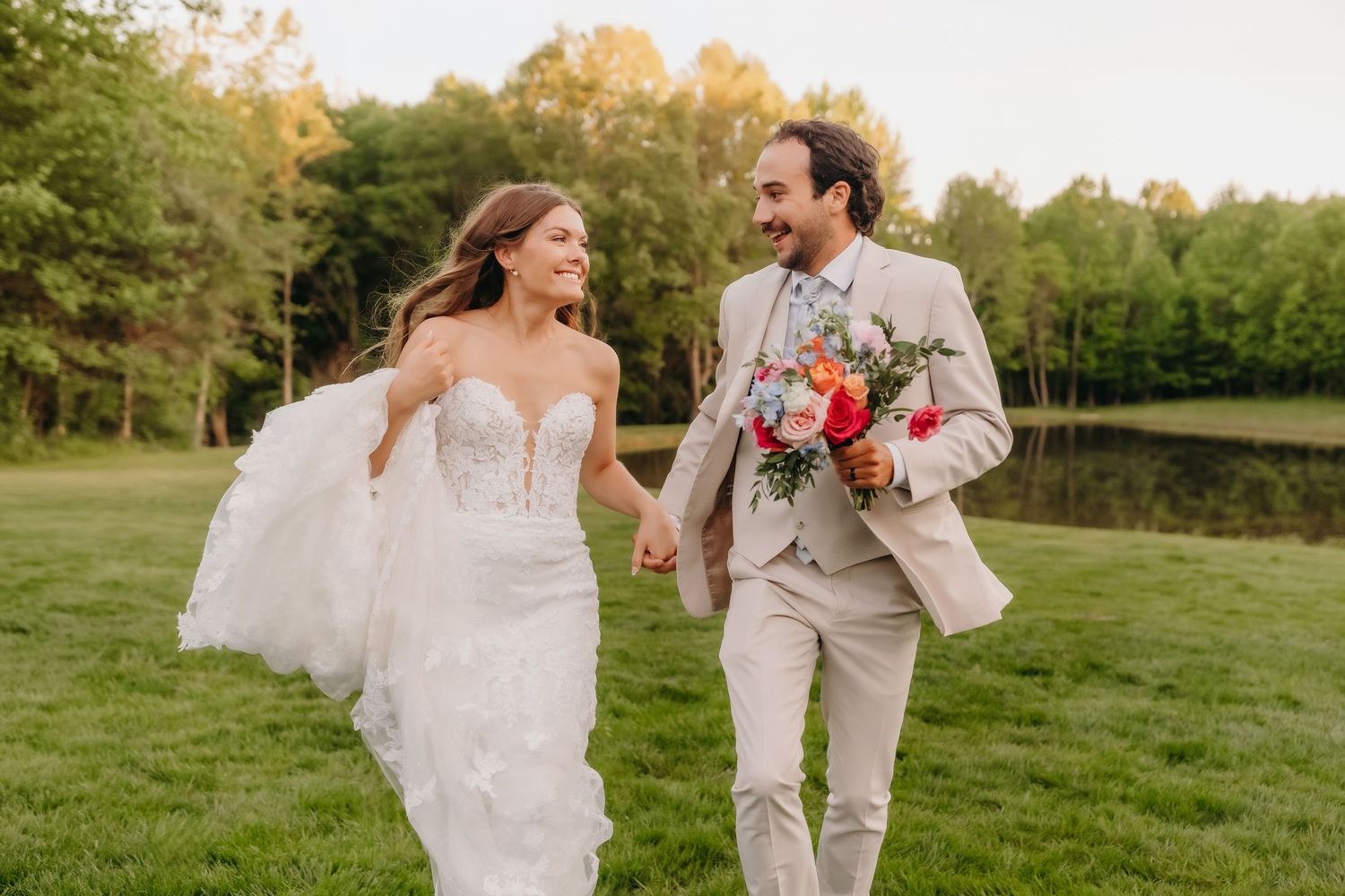 Bride in lace gown and groom in tan suit walk hand in hand across a green lawn at sunset.