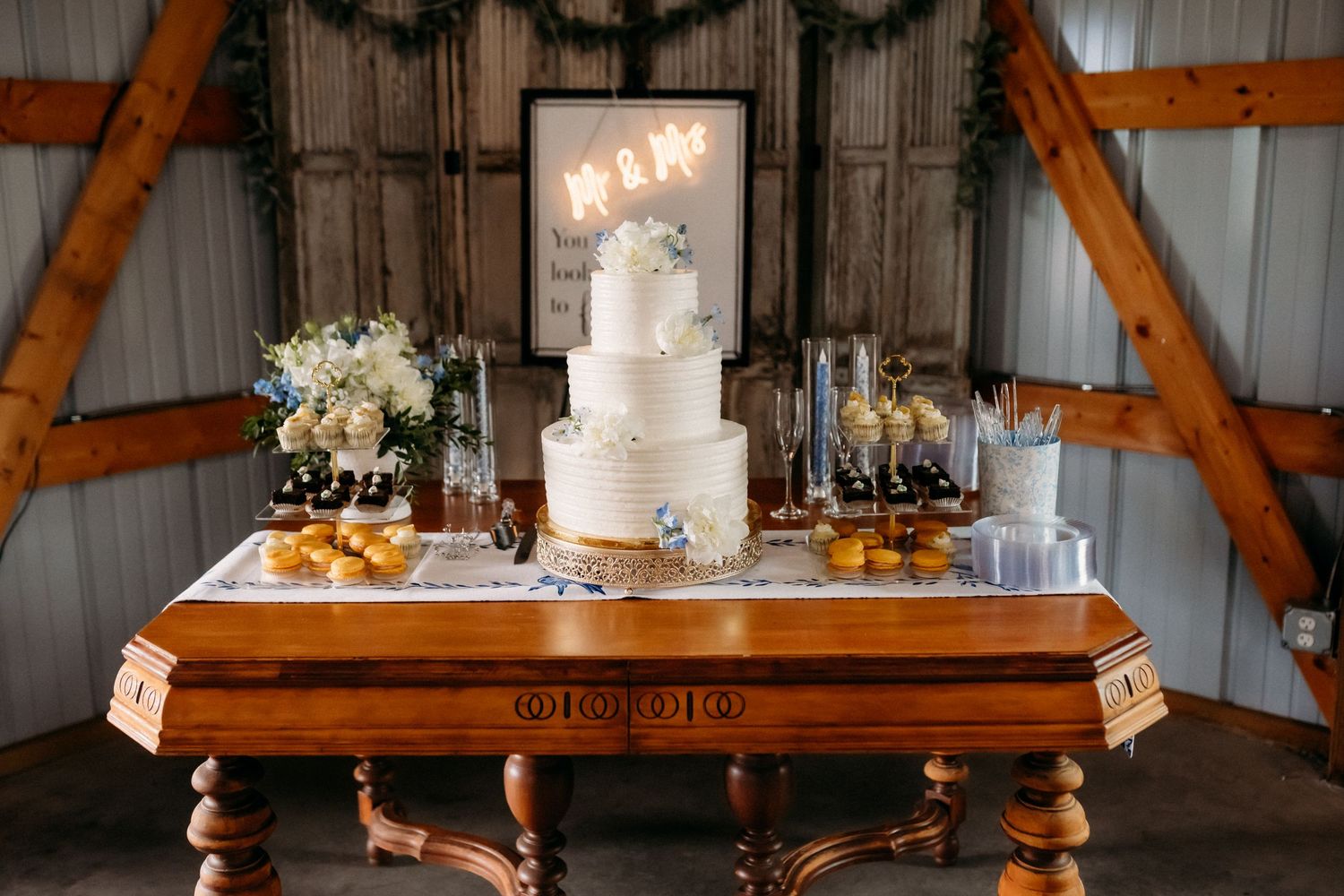 Elegant three-tier white wedding cake displayed on a wooden table with floral decor in a rustic barn venue.