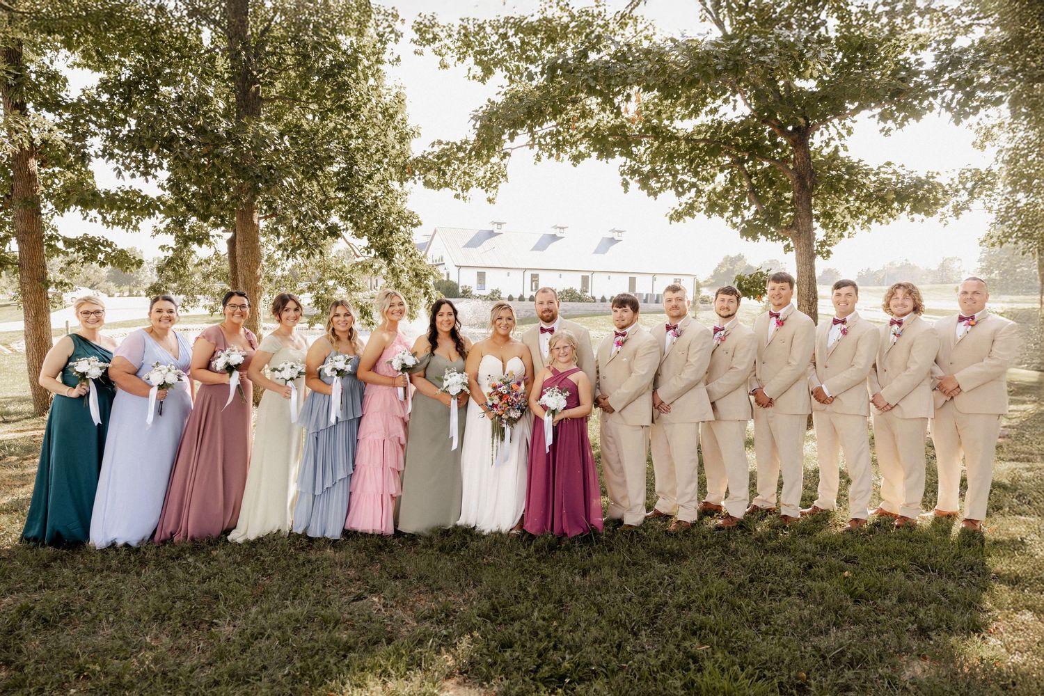Wedding party in pastel colored dresses lined up outdoors at Phillips Grove under trees at sunset.