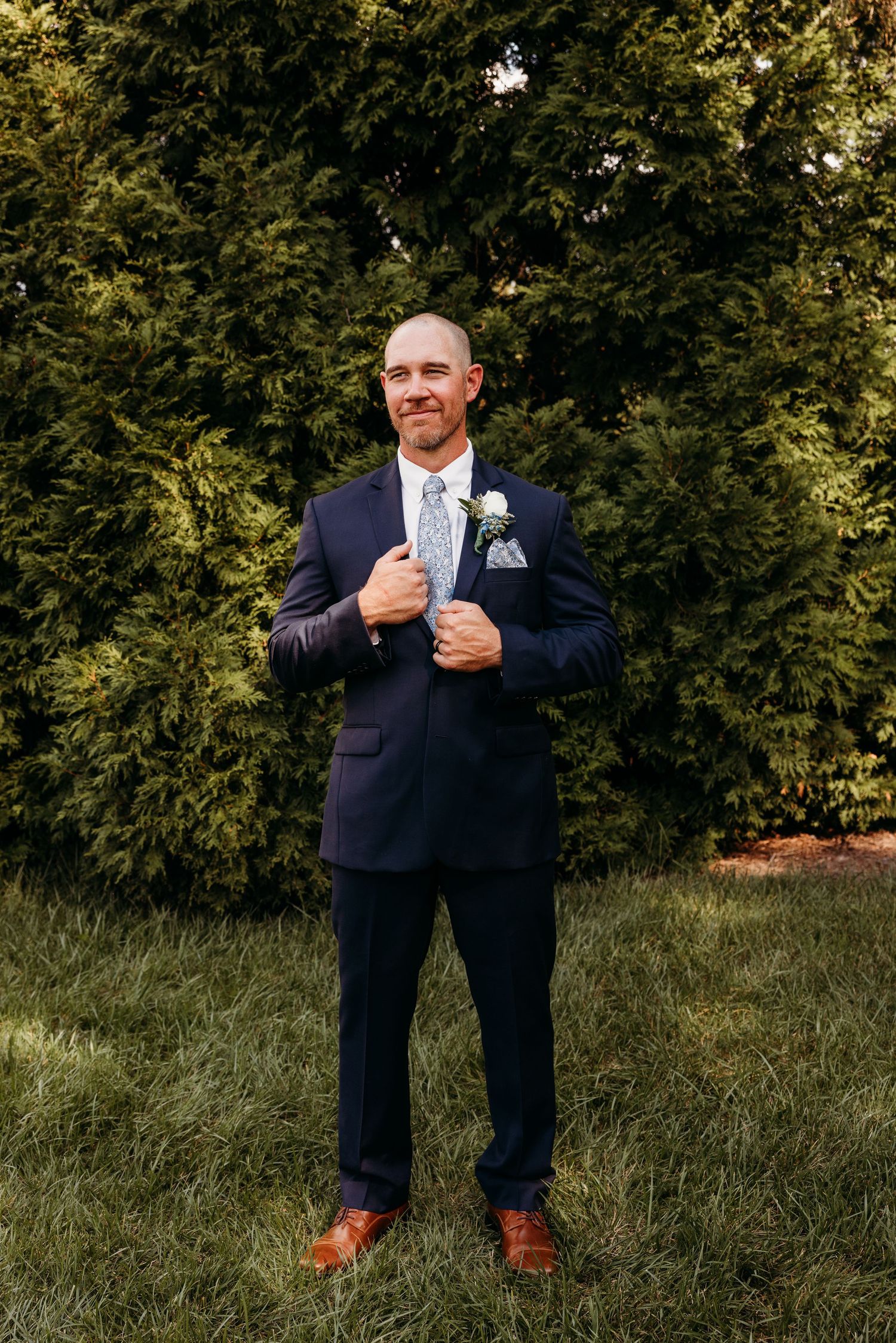 Groom adjusting suit jacket while standing in grassy outdoor setting.