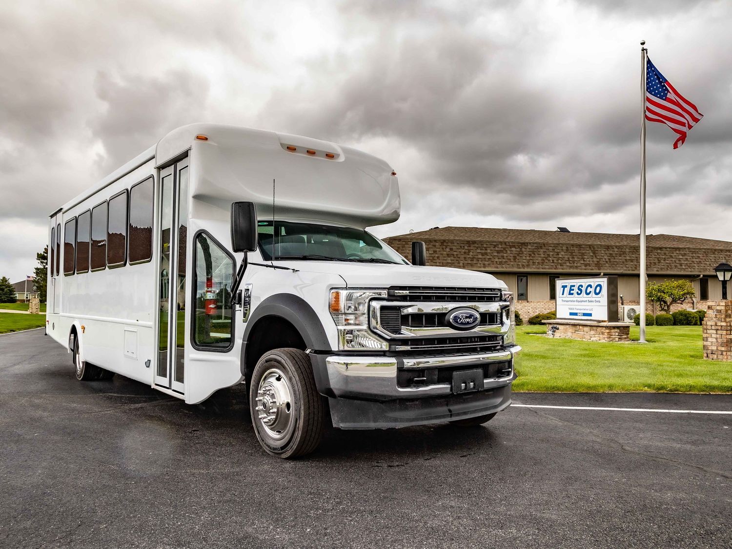 White school bus parked in front of a brick building with American flag under cloudy skies.