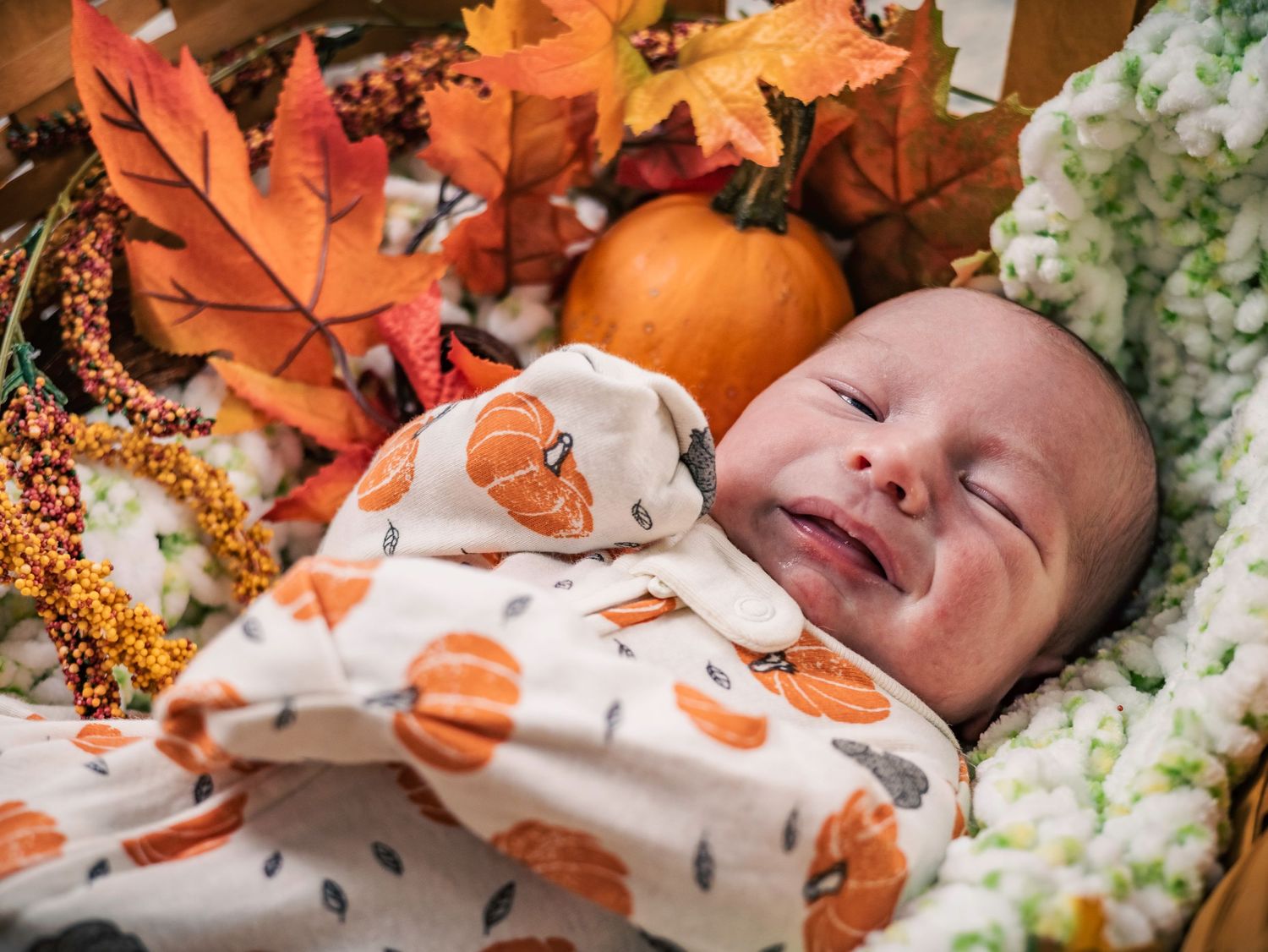 Baby in pumpkin-print outfit smiling among fall decorations and orange leaves.