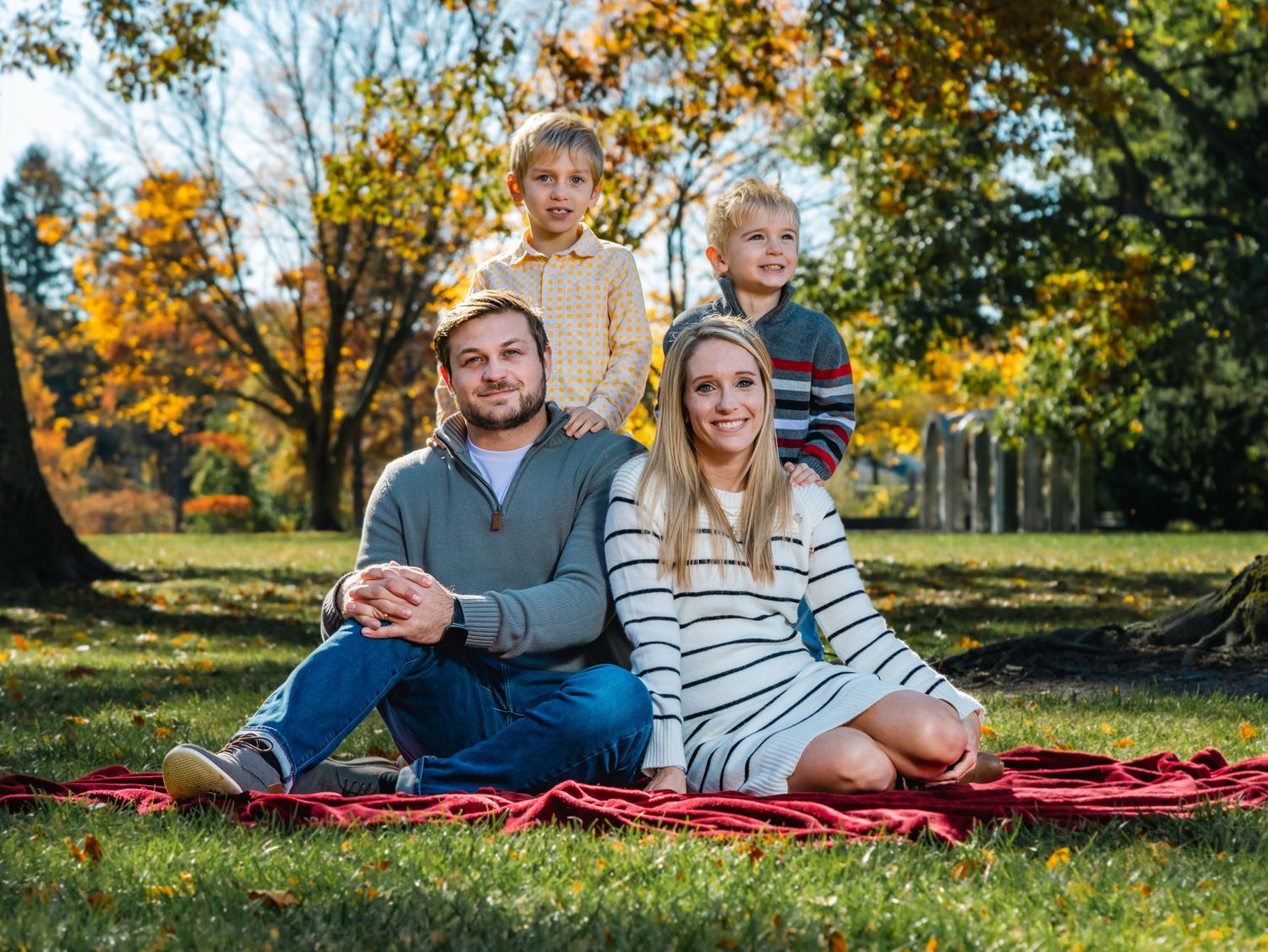 Family enjoying autumn picnic outdoors on red blanket surrounded by fall foliage.
