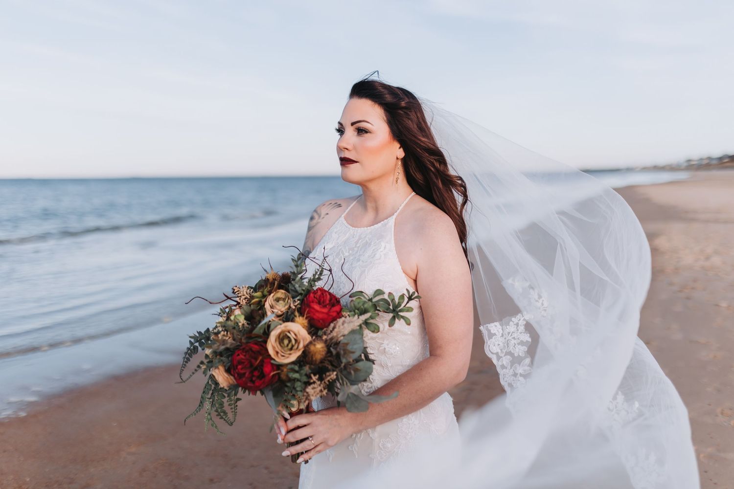 A person in a flowing white wedding dress holds a colorful bouquet while walking along a sandy beach at sunset.