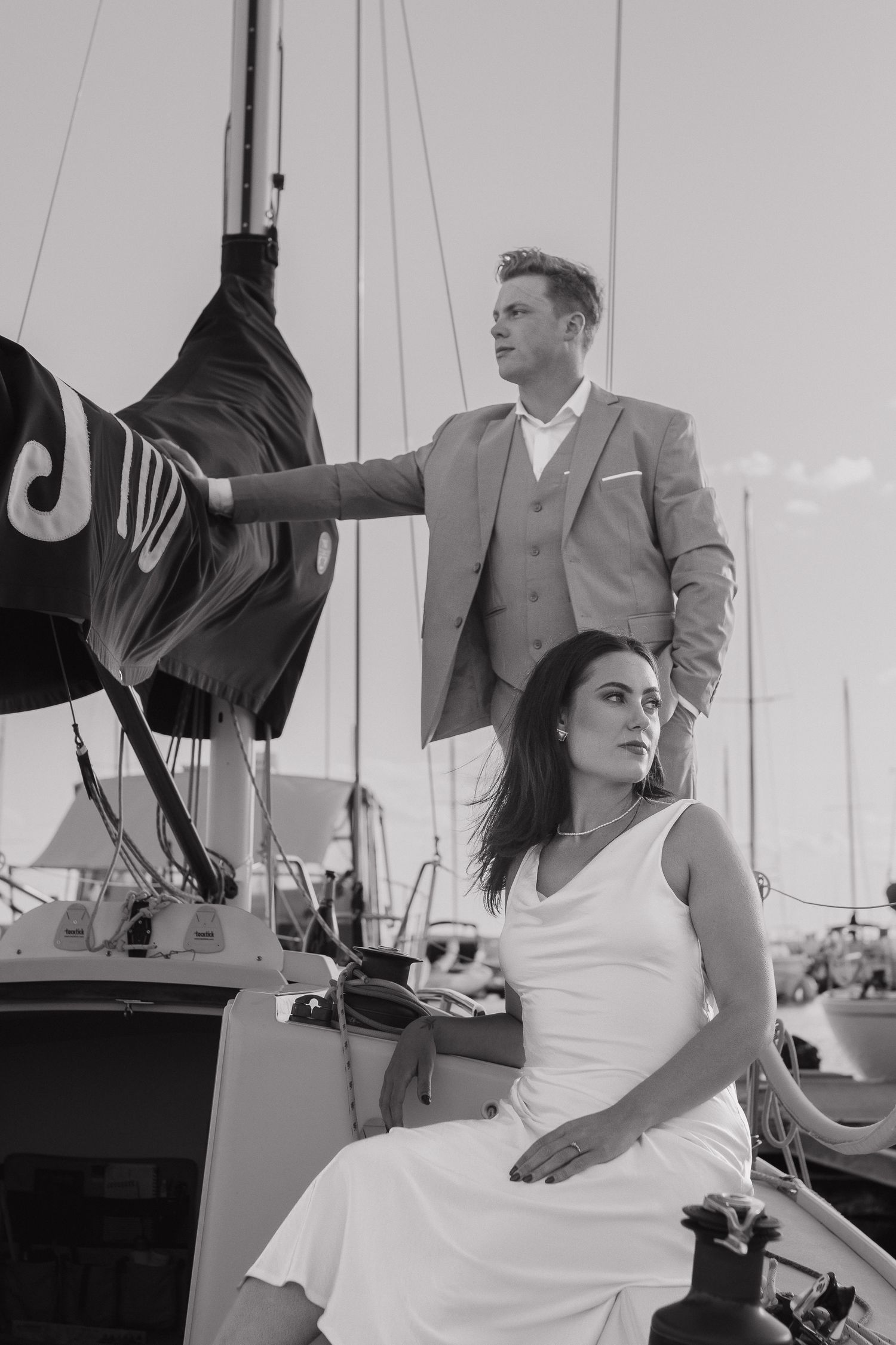 A black and white photo of a couple in elegant attire aboard a sailboat at a marina.