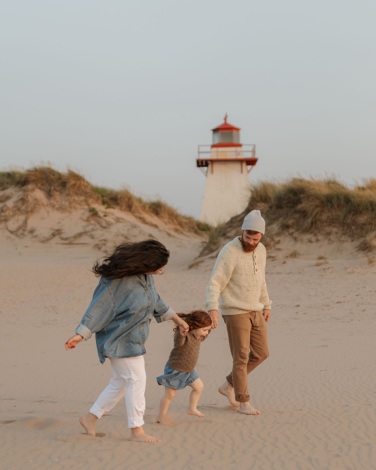 Family strolling on sandy beach near red and white lighthouse during golden hour.