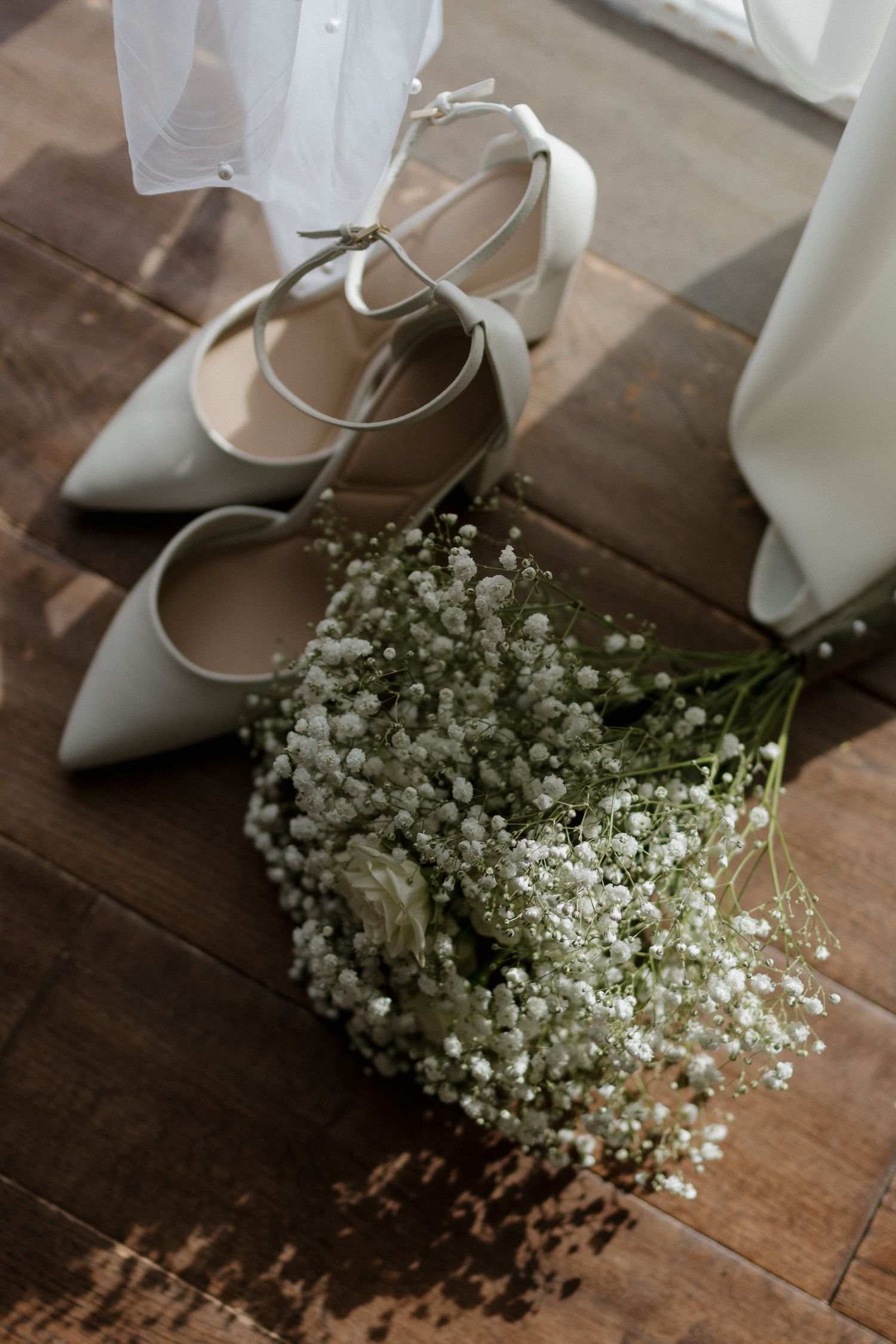 Elegant white bridal shoes with a delicate baby's breath bouquet on wooden flooring.