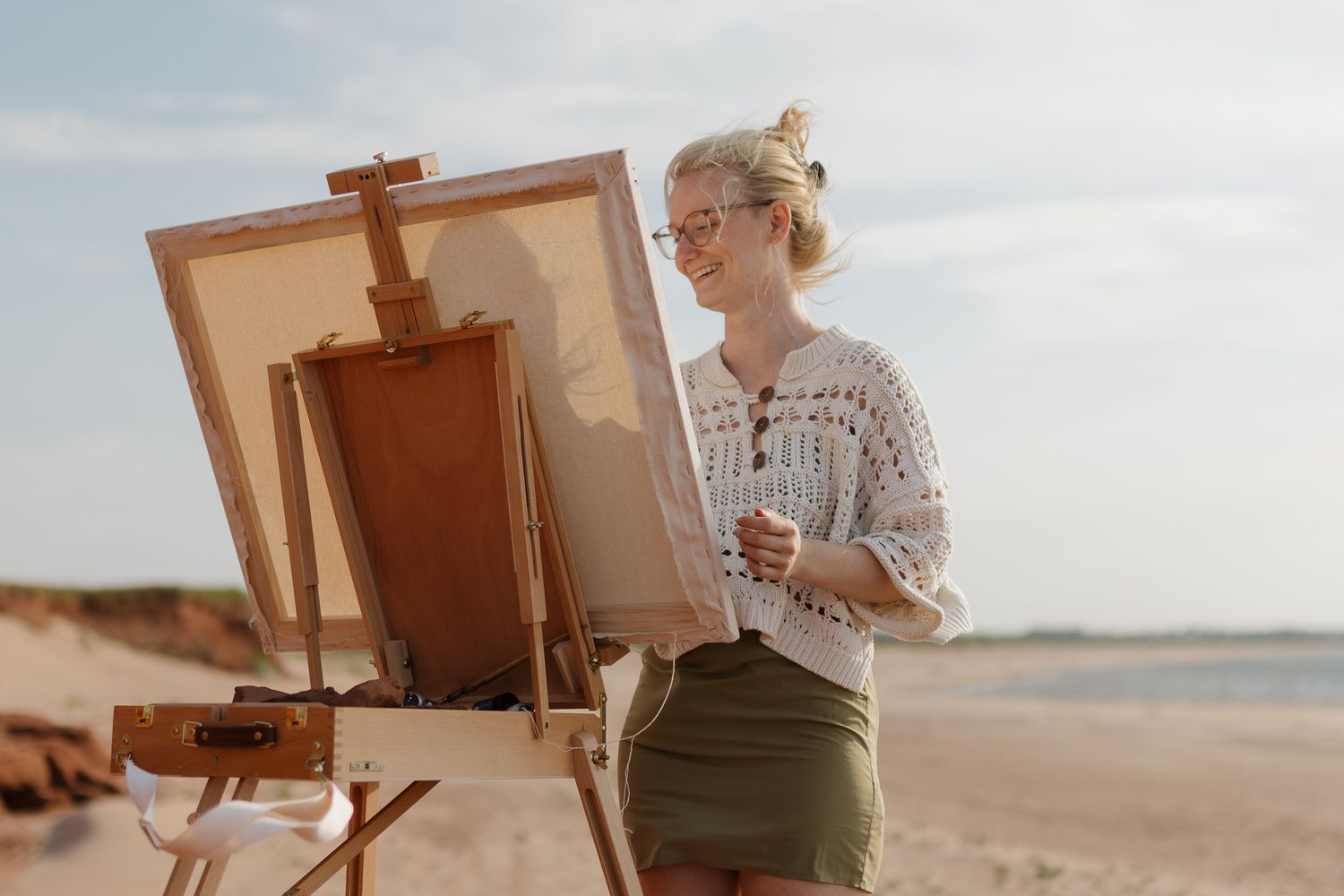 Artist painting at easel on beach with sun setting in background.