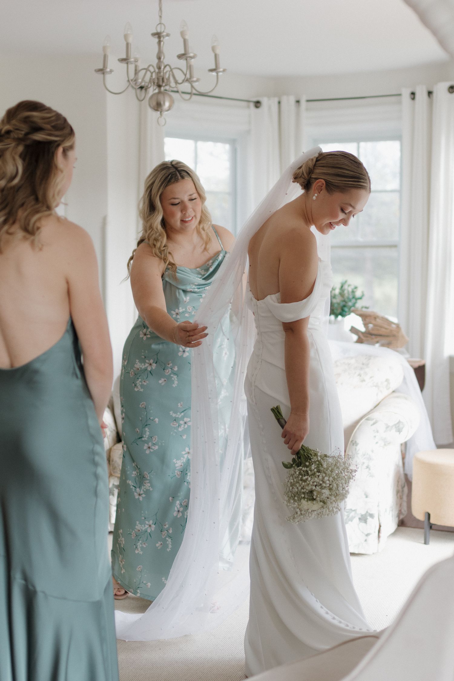 Women in teal bridesmaid dresses help with wedding preparations in bright room.