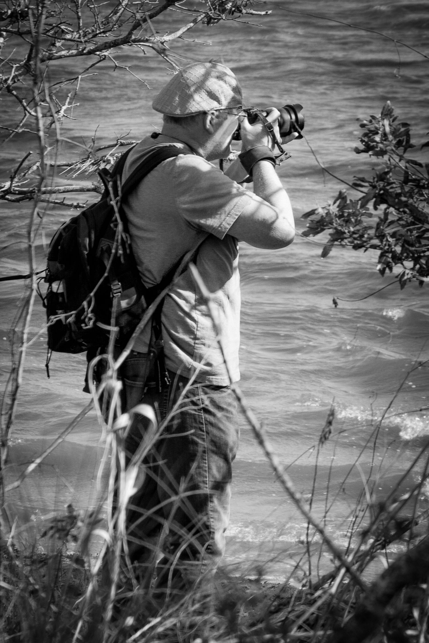 Photographer Ed Hineline capturing nature along a shoreline, framed in black-and-white with camera and backpack.