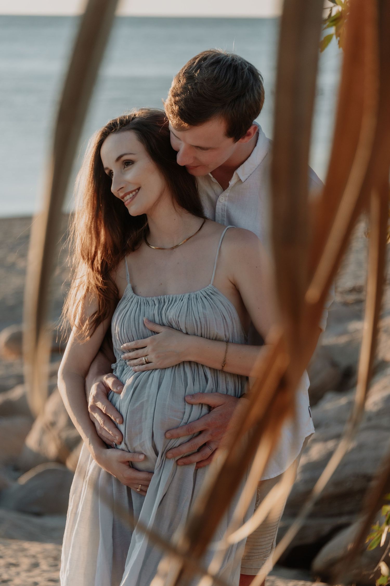 A romantic maternity photoshoot on the beach at sunset with a light blue flowy dress and natural elements framing the scene.