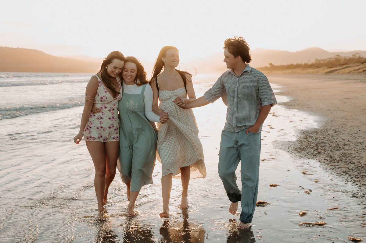 Four people walking and laughing together on a sunny beach at golden hour with waves and mountains in the background.