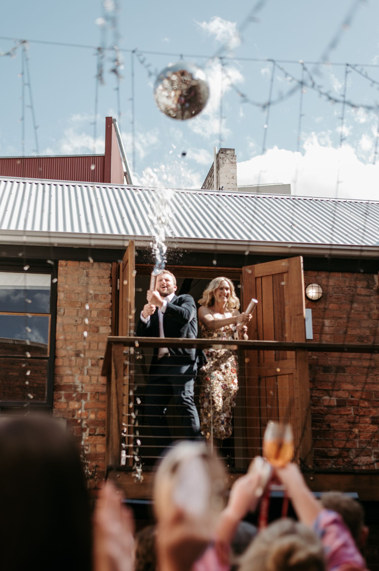 Couple celebrates wedding with champagne spray and confetti on a rustic brick venue balcony under sunny skies.