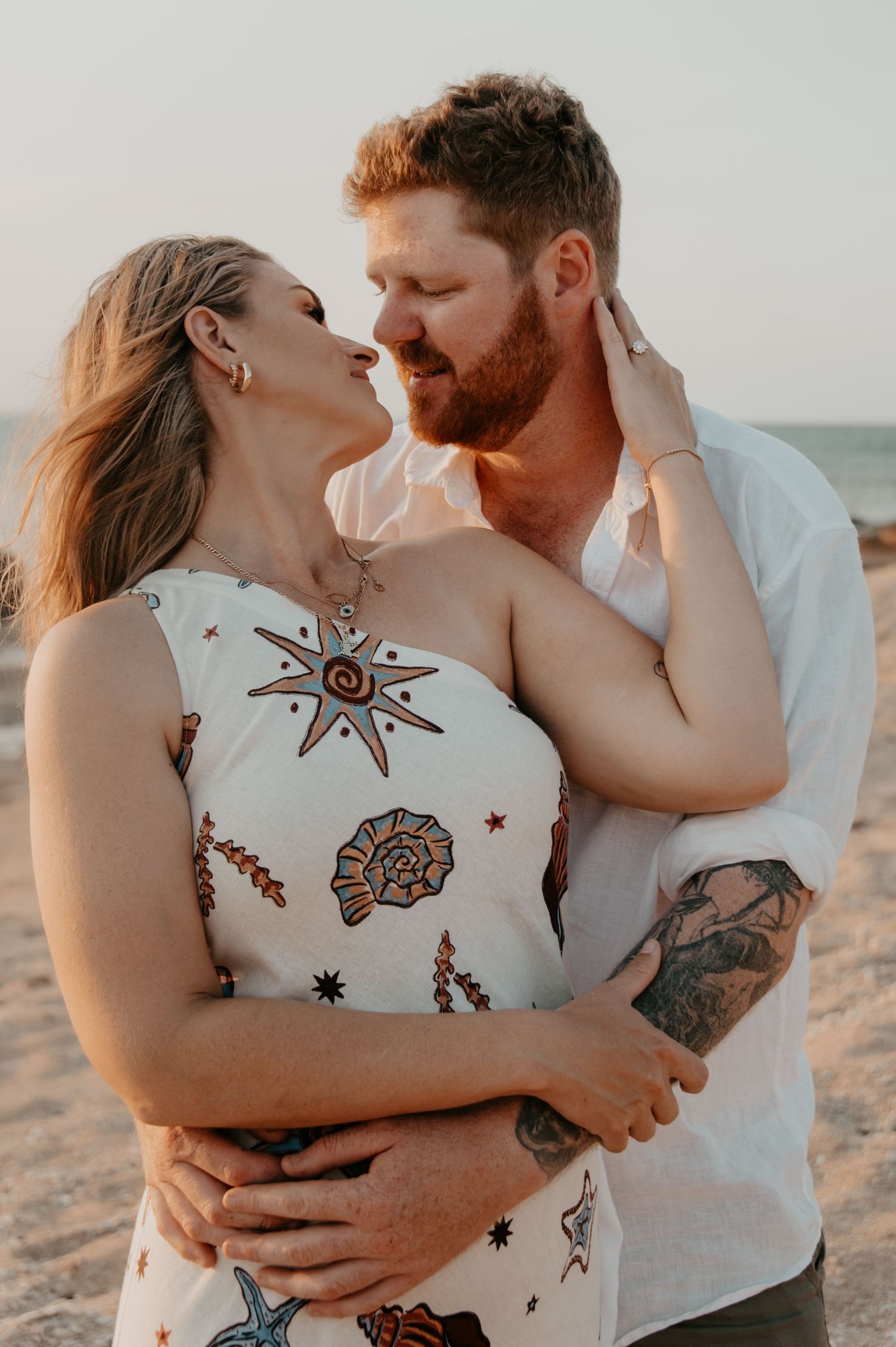 Romantic couple embraces at sunset on beach, woman in celestial print dress and man in white shirt.