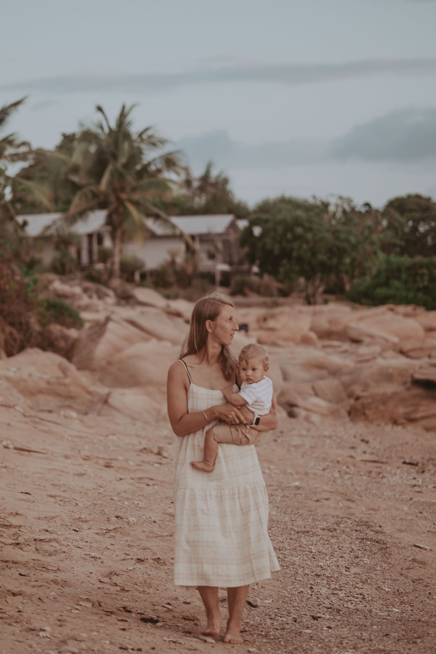 Woman in white dress holds baby while standing on reddish rocky terrain with palm trees and buildings in background.