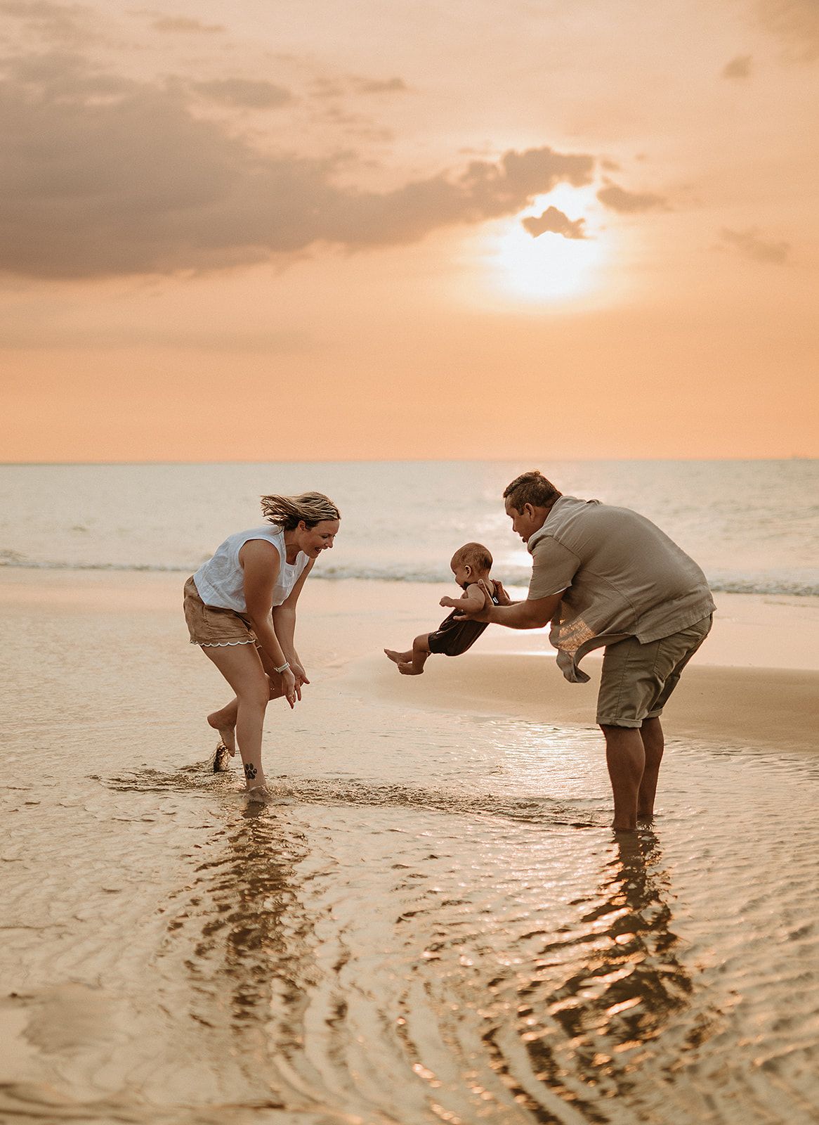 Family plays together at sunset on a beach, splashing in shallow water with golden sky above.