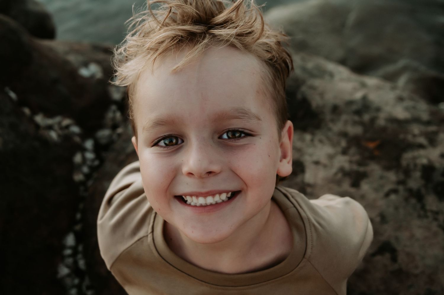 Close up portrait of a child with blue eyes wearing a tan shirt and smiling outdoors.
