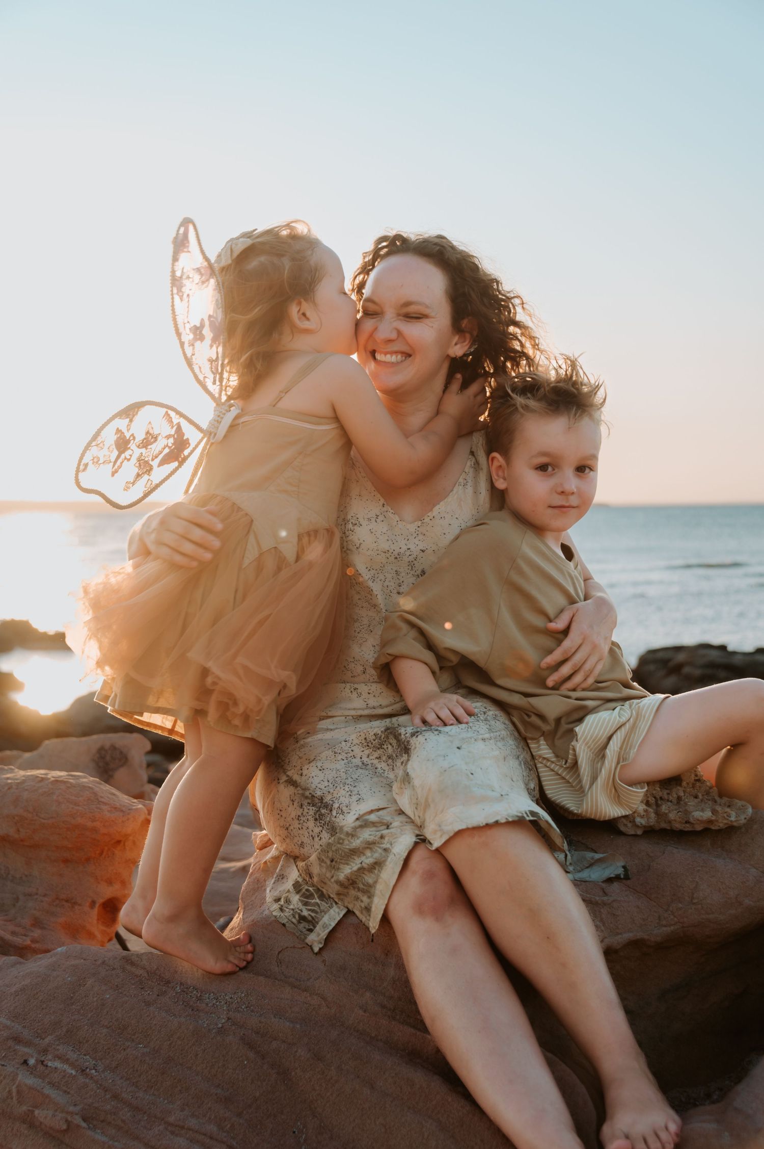 A joyful family moment captured at sunset on rocky coastal beach with warm golden light filtering through.