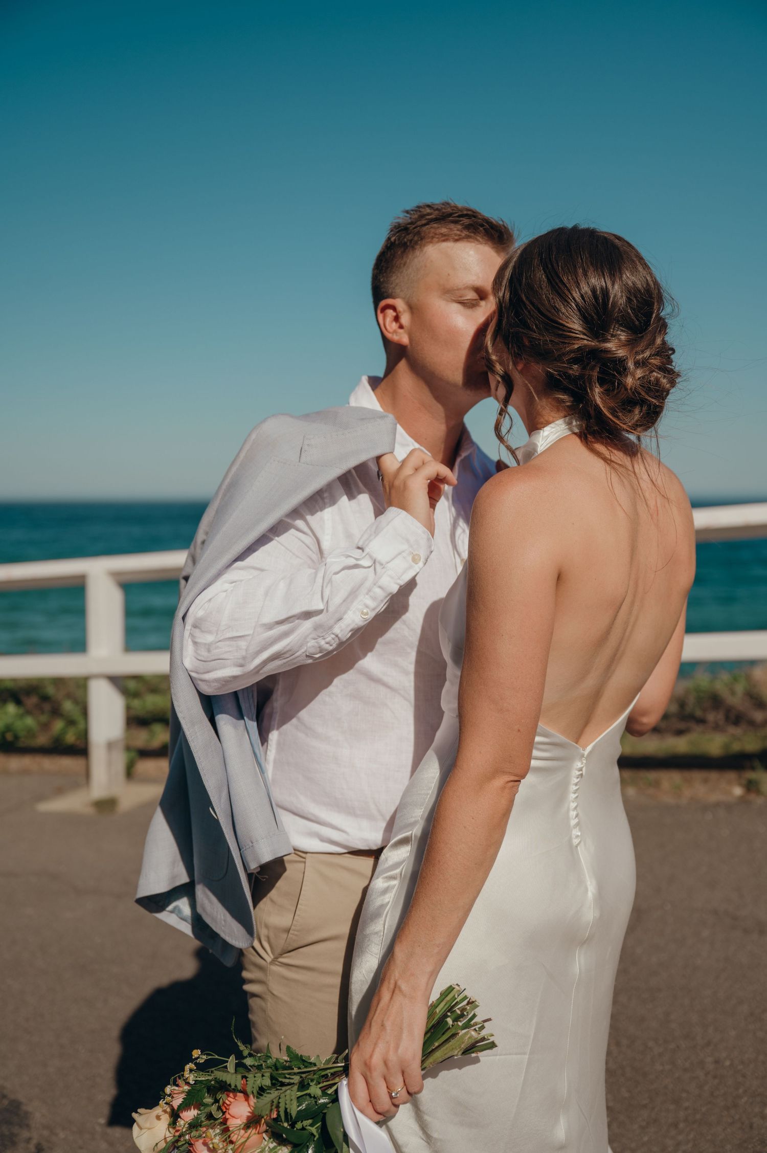 Bride and groom share a kiss outdoors by the ocean, she holds a bouquet in a backless white gown.
