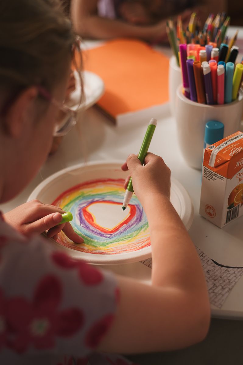 A young girl draws a colorful rainbow heart on a paper plate using crayons at a craft table.