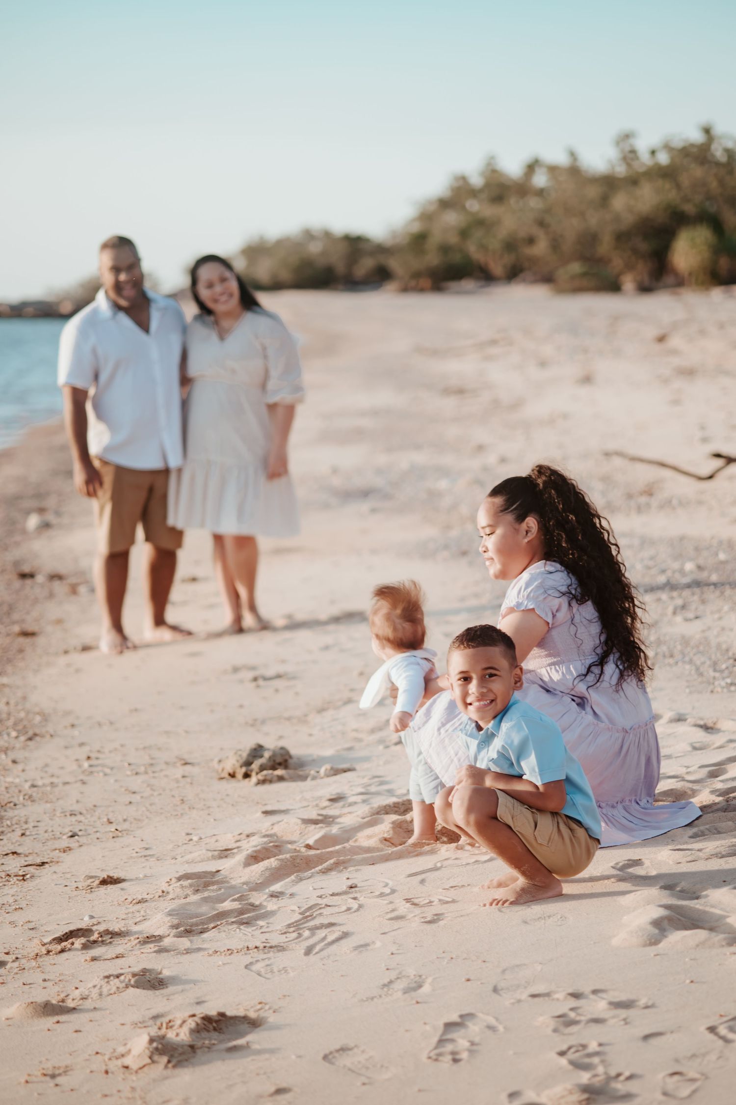A family spending time together on a sandy beach during golden hour with ocean and trees in the background.