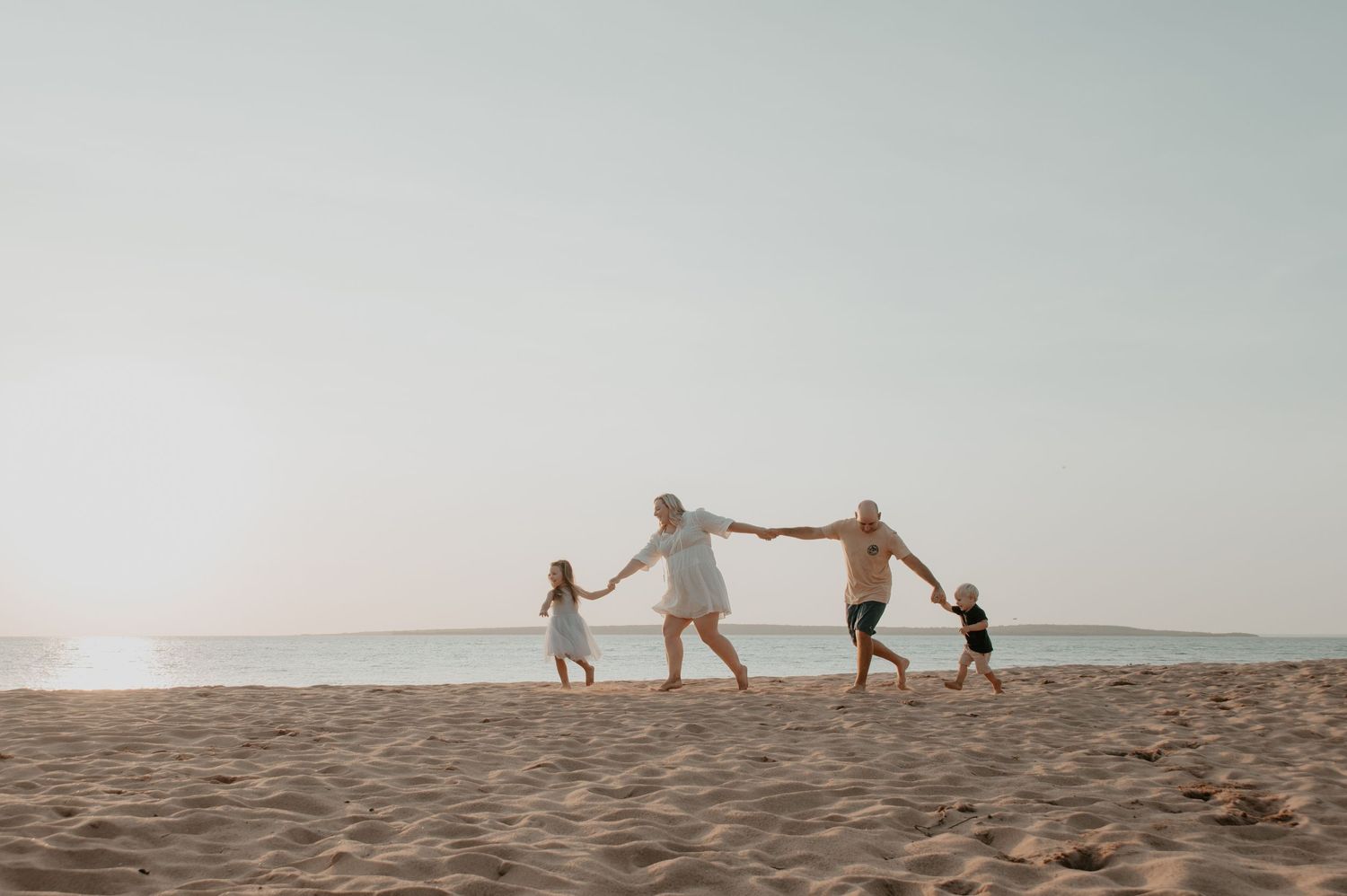 Group holds hands and runs together on a sandy beach at sunset silhouetted against the bright sky.