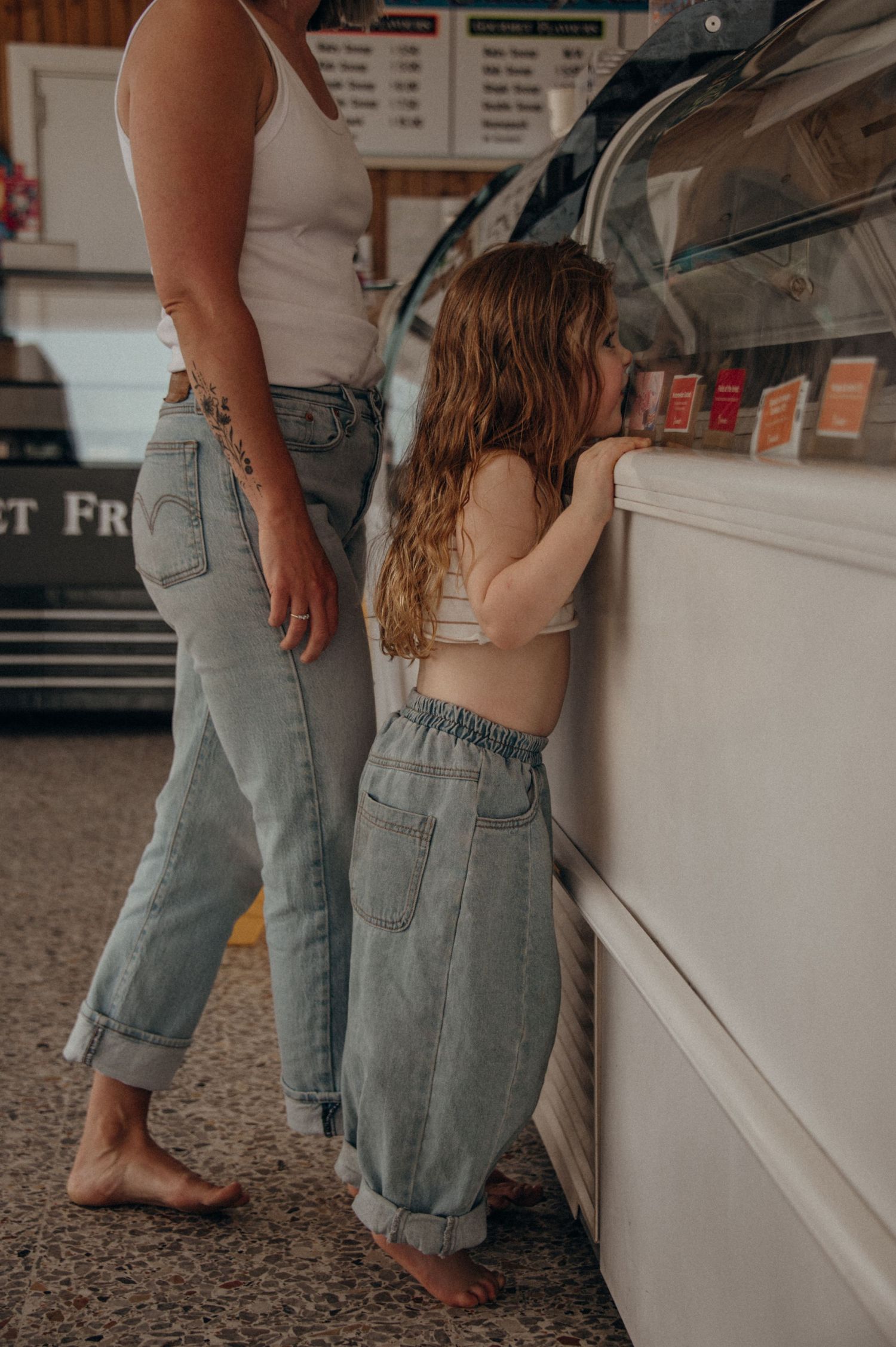 Two barefoot people in rolled up jeans standing at an ice cream counter looking at dessert selections.