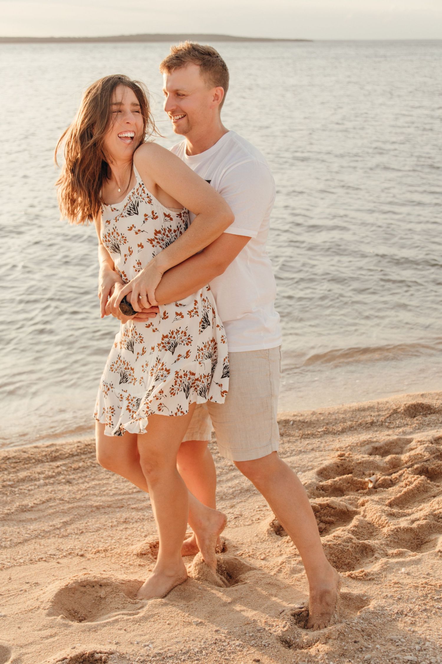 Couple embracing on a beach at sunset, woman in floral dress laughing as man hugs her from behind, ocean in background.