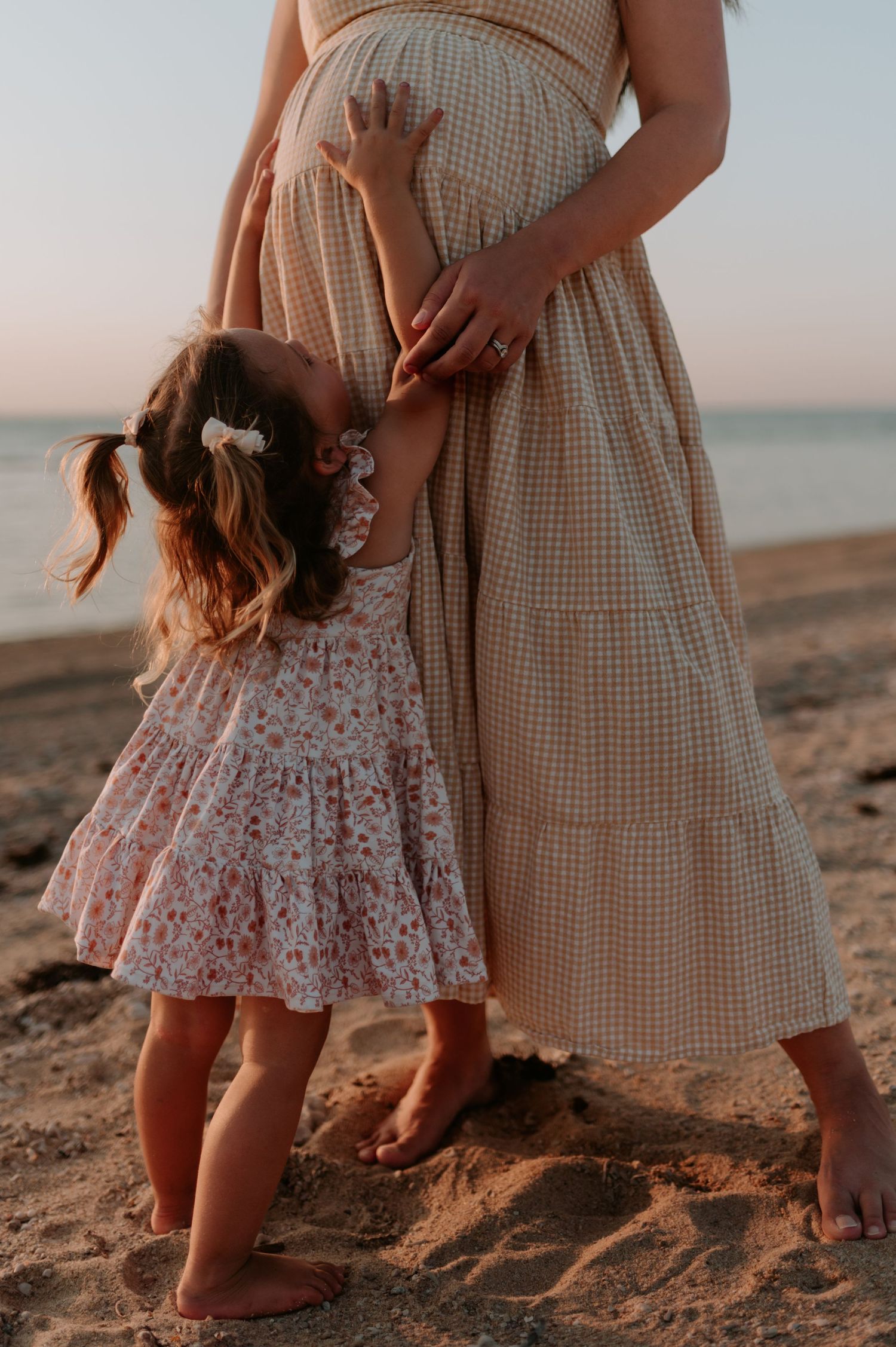 Parent and child in flowing dresses share tender moment on sandy beach at sunset.
