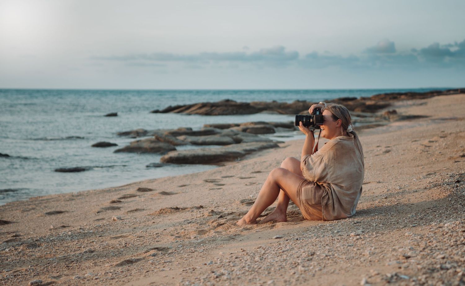 Someone sits on a rocky beach shore in casual clothing, looking out at the ocean on a serene evening.