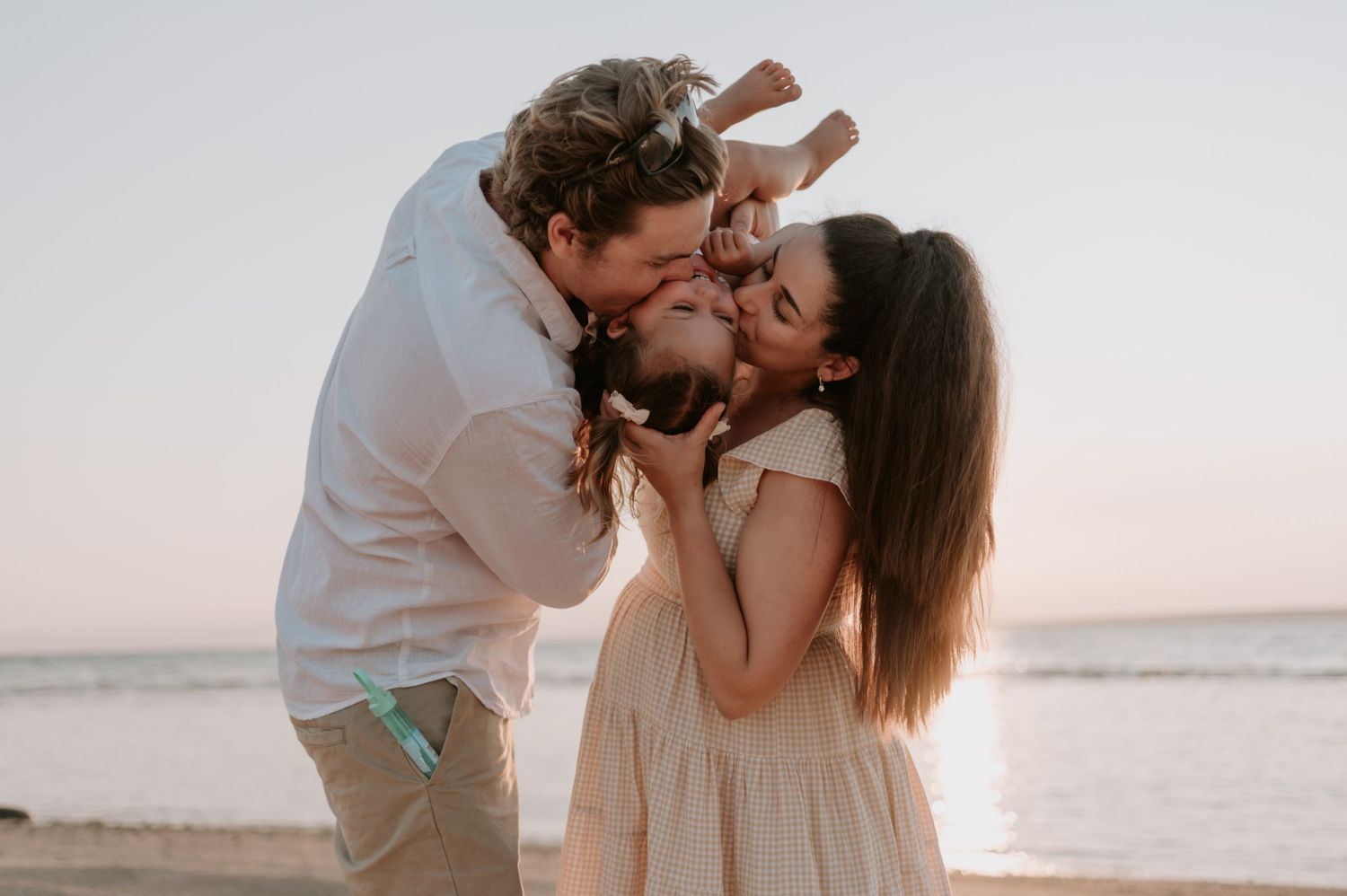 A romantic couple embraces and kisses on the beach at sunset, with the ocean visible in the background.