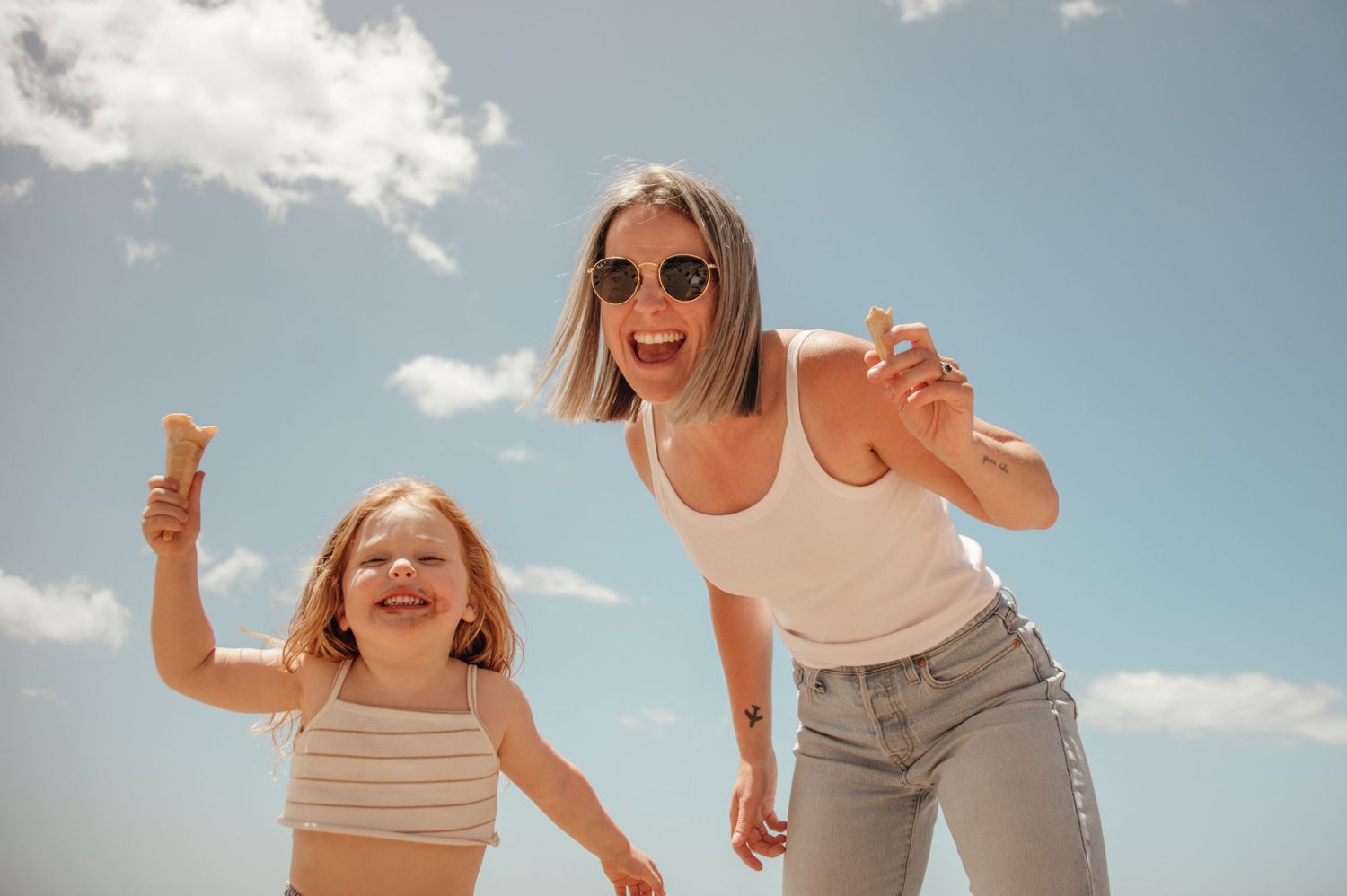 Two people in summer outfits hold ice cream cones while jumping joyfully against a bright blue sky with white clouds.