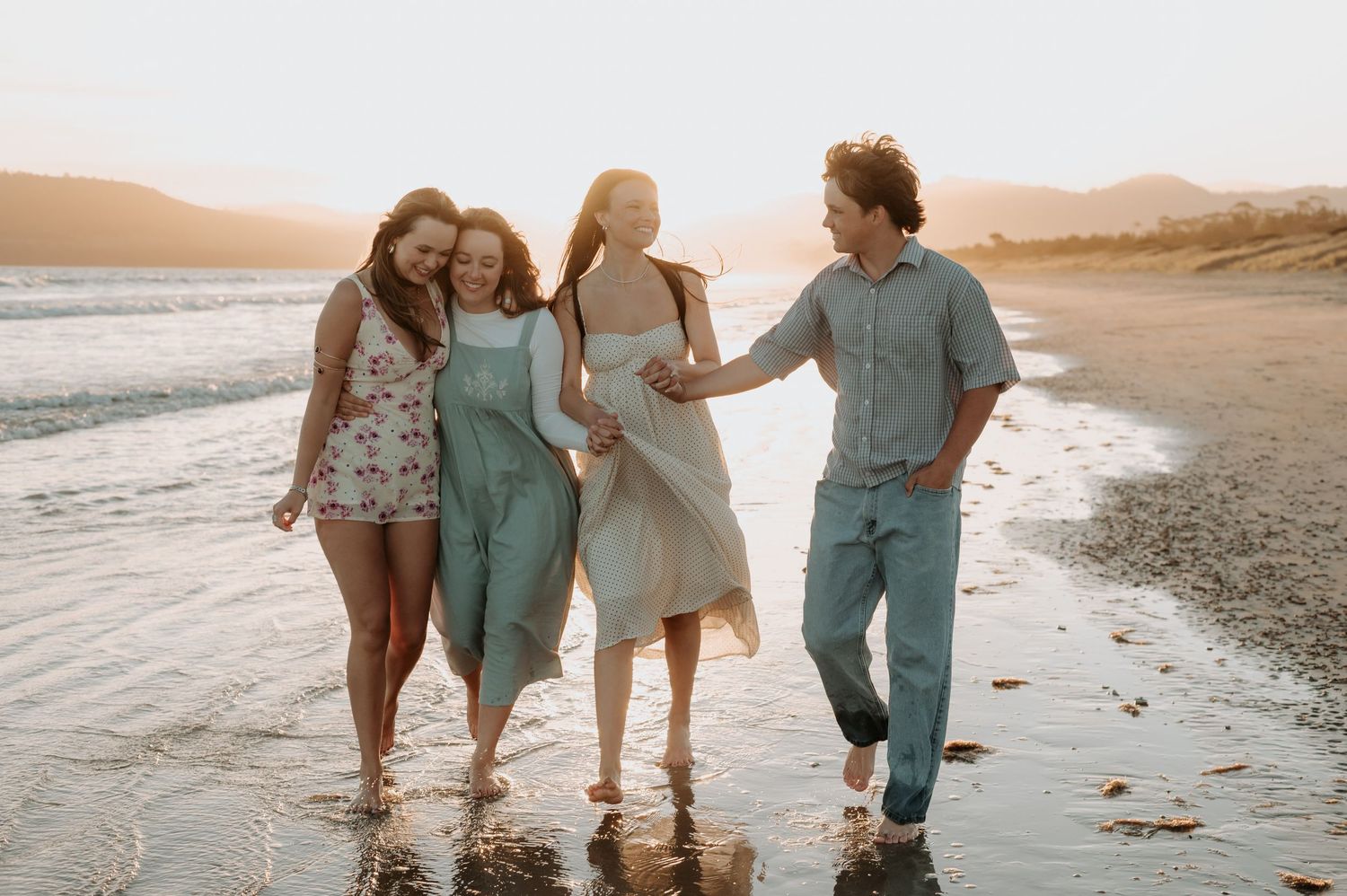 Four friends walking and laughing together on a sunny beach at golden hour with mountains in the background.