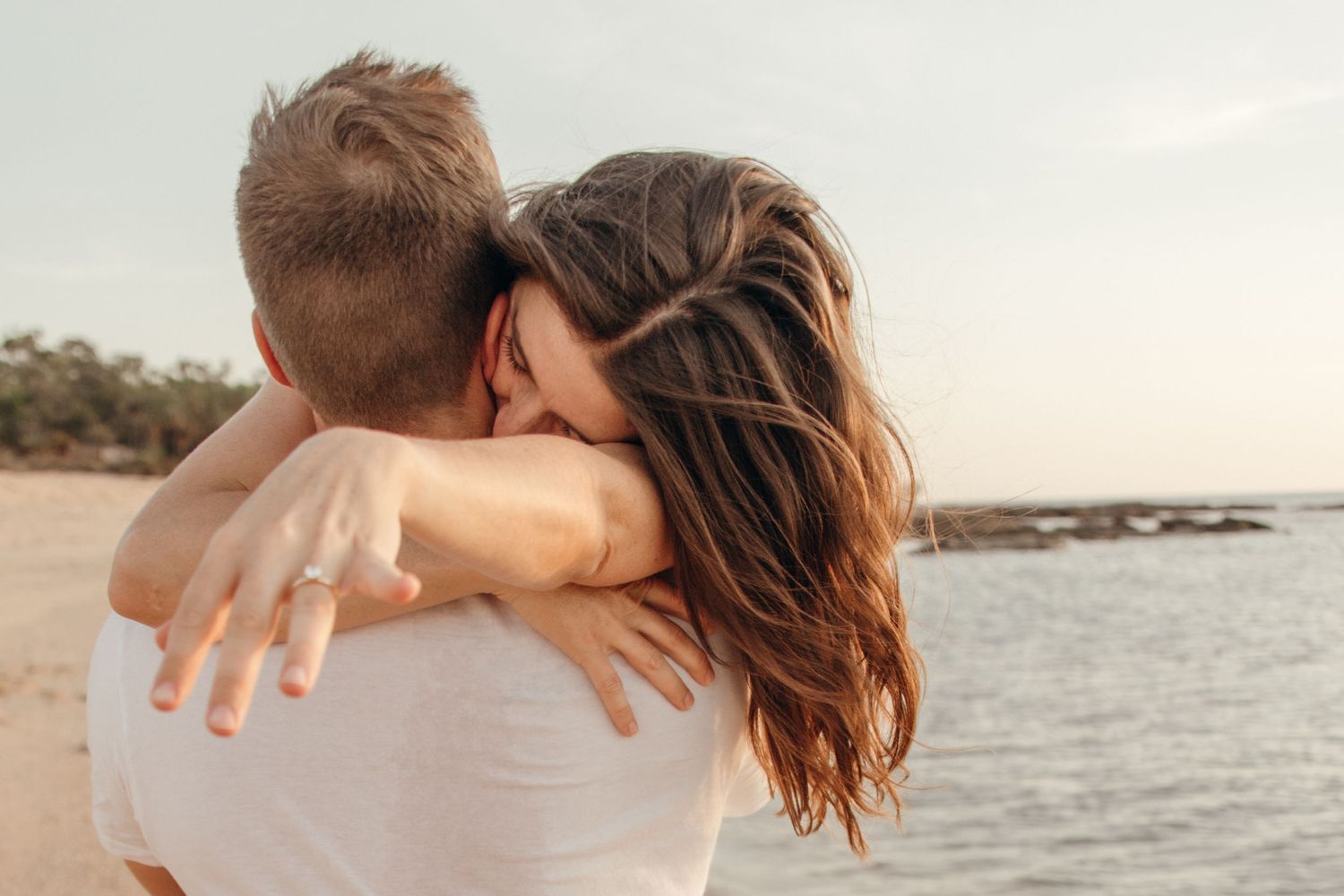 Couple embraces on beach at sunset with ocean waves and rocky coastline in background.